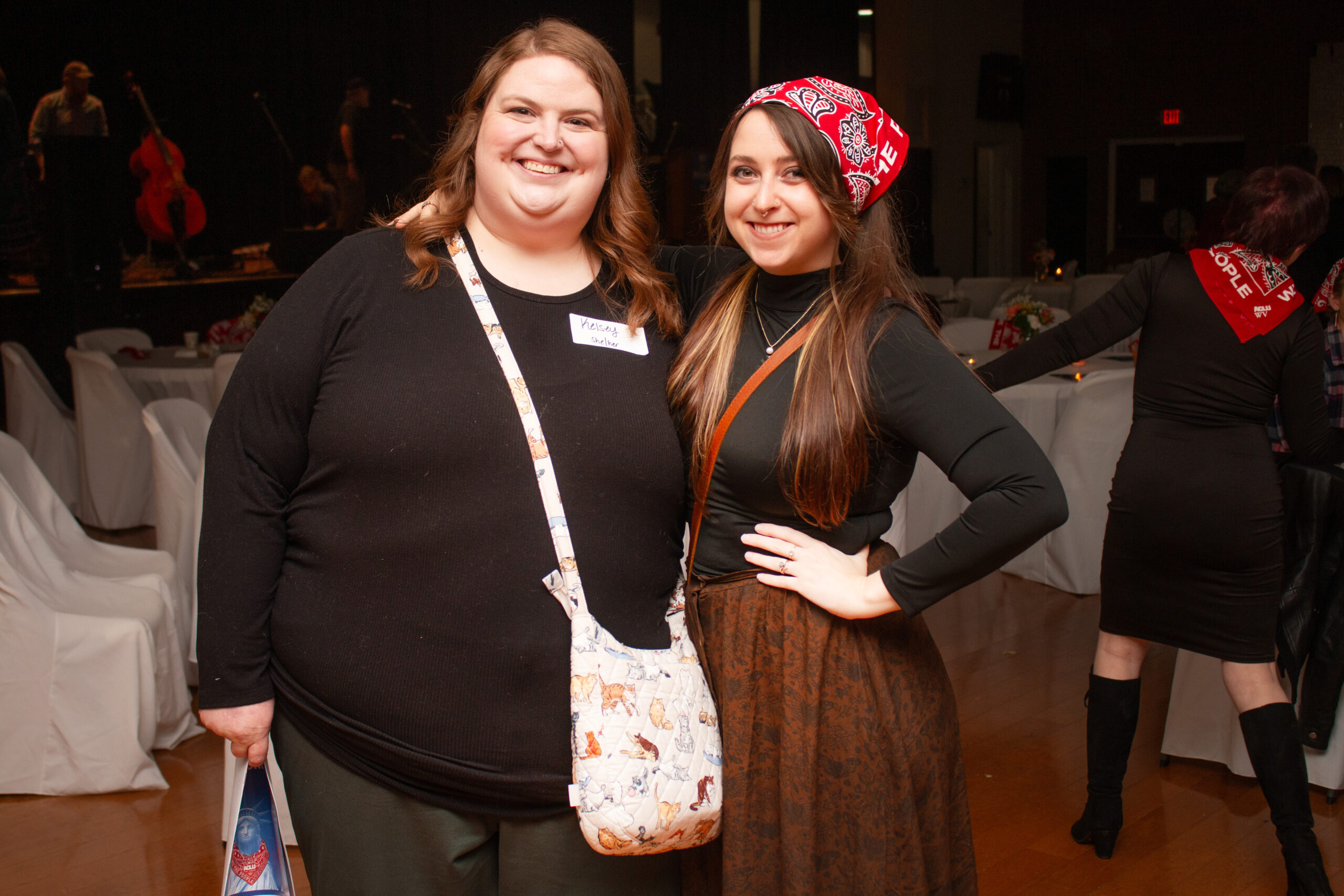 Two people stand together smiling at an evening event venue with white-covered chairs and a stage visible in the background. The person on left wears all black with a crossbody bag, while the person on right wears black long sleeves with a brown patterned skirt and a red bandana headband.