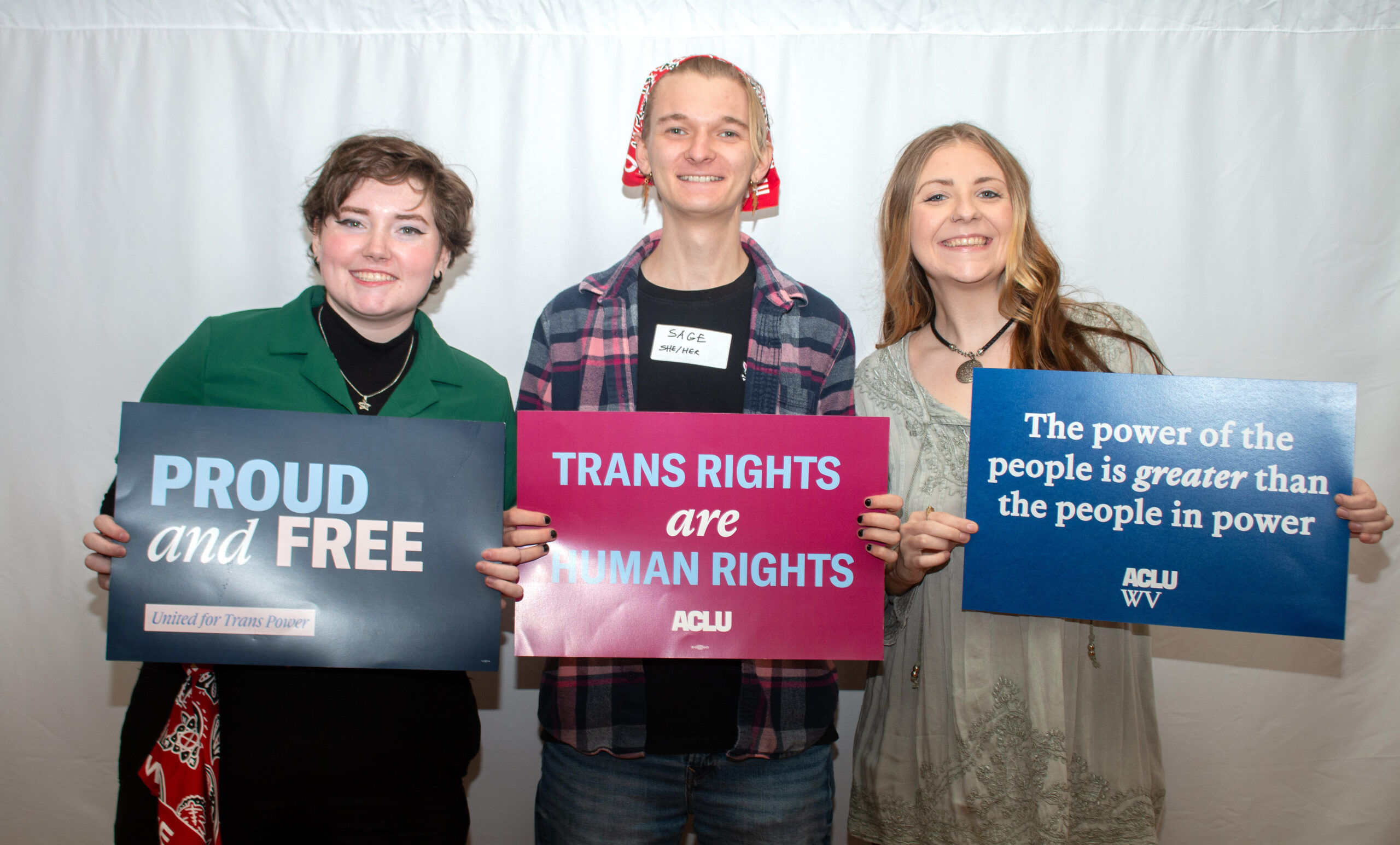 Three people pose together holding advocacy signs against a white backdrop. From left: person in green jacket with "PROUD and FREE" sign, person in center wearing plaid flannel and red bandana headwear holding pink "TRANS RIGHTS are HUMAN RIGHTS" ACLU sign, and person on right in gray top holding blue ACLU WV sign reading "The power of the people is greater than the people in power.