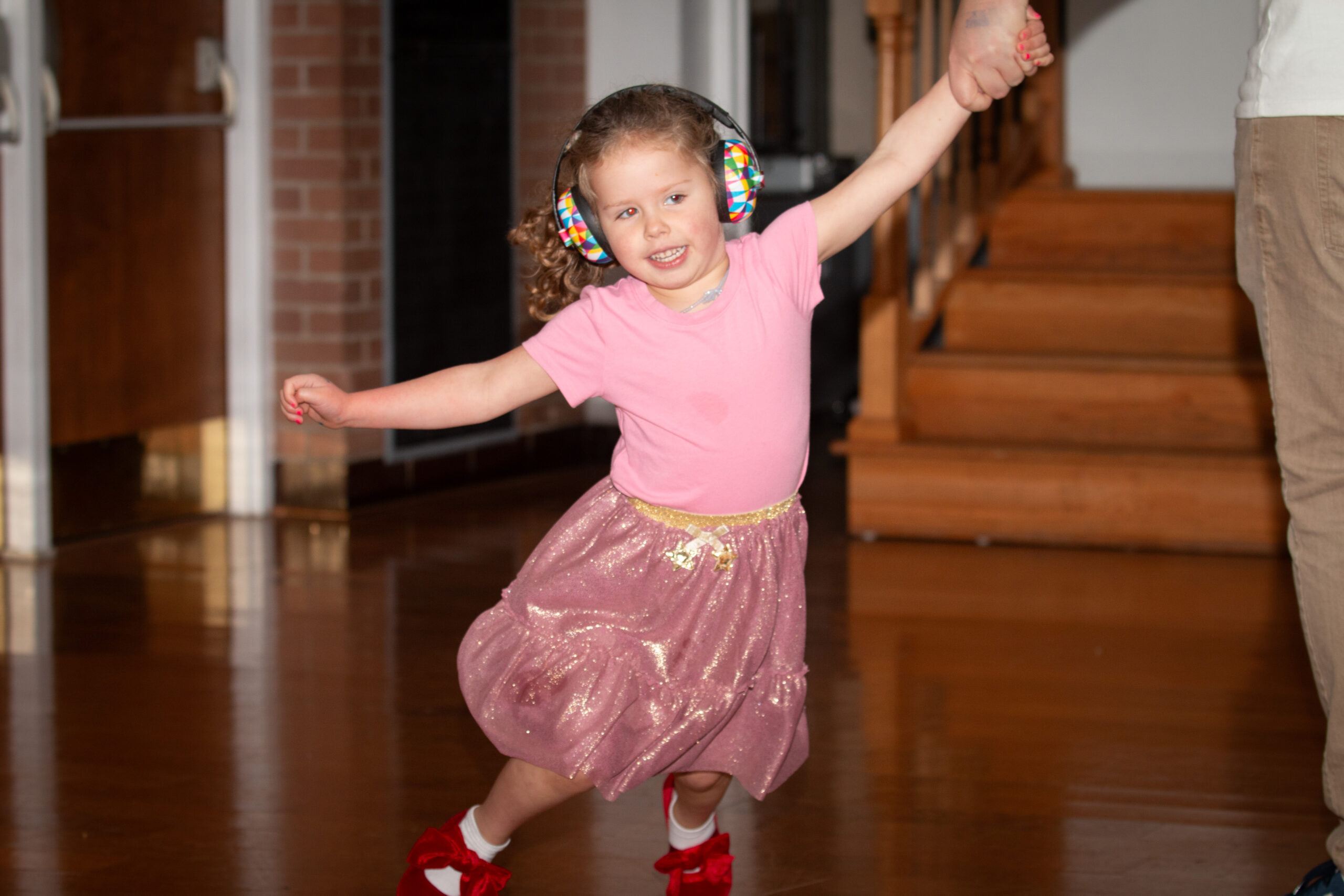 A young child with curly hair wearing colorful checkered ear protection, a pink t-shirt, and a sparkly pink skirt dances joyfully with both arms raised. The setting appears to be an indoor venue with wooden architectural elements visible in the background.Retry