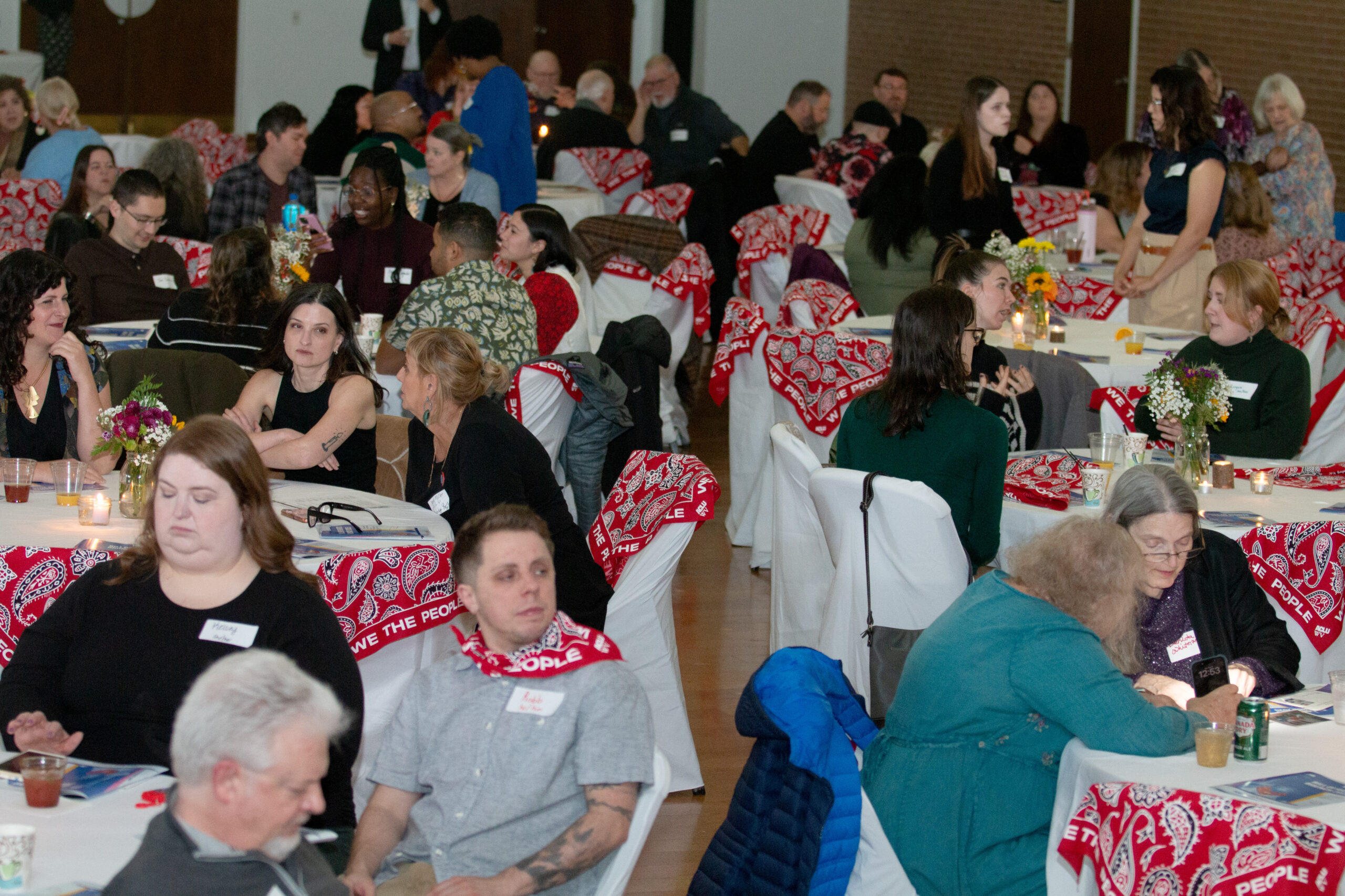 A crowded event hall filled with attendees seated at round tables covered with white tablecloths. Red bandanas with "WE THE PEOPLE" text are placed on chairs throughout the space. Tables feature small floral centerpieces with candles. The diverse crowd mingles and converses in an atmosphere decorated with string lights overhead.