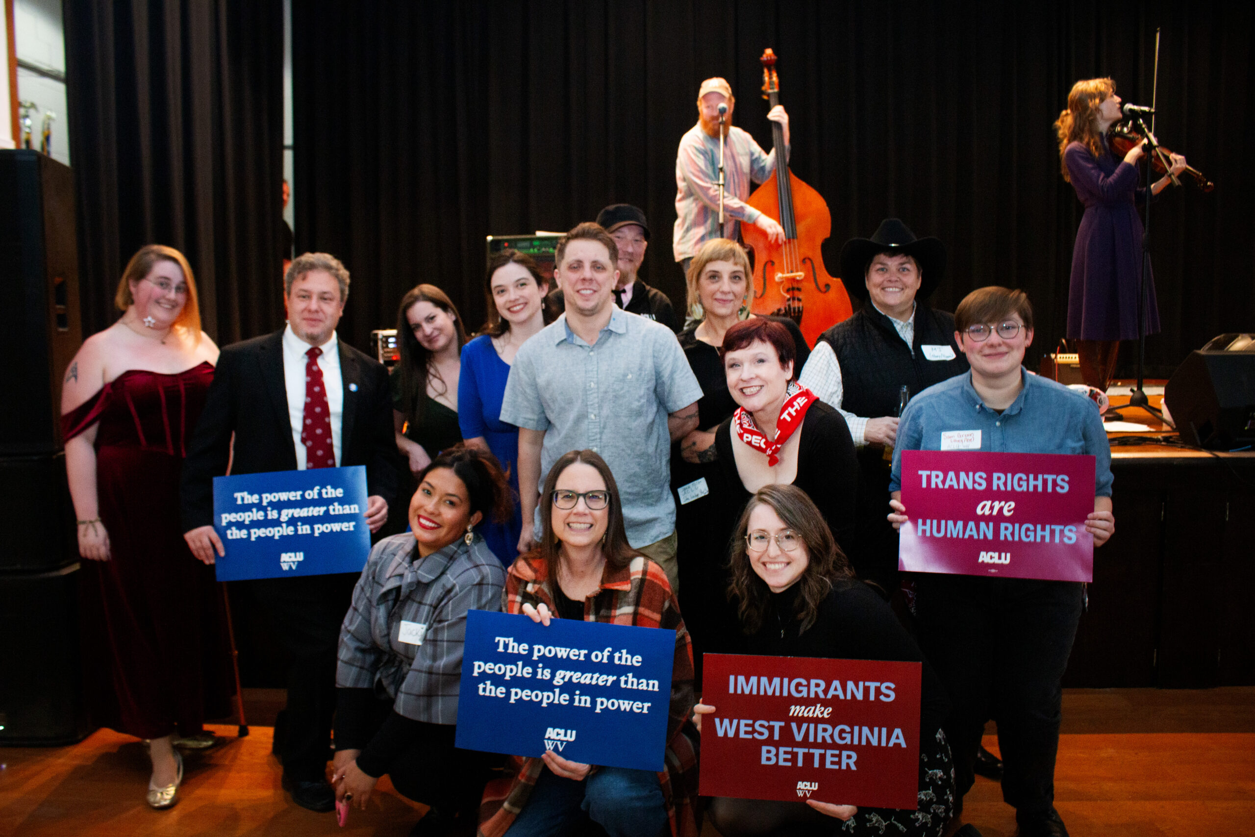 A large group poses on stage with musicians visible in the background playing upright bass and fiddle. The group holds various ACLU advocacy signs including "The power of the people is greater than the people in power," "TRANS RIGHTS are HUMAN RIGHTS," and "IMMIGRANTS make WEST VIRGINIA BETTER." People wear formal and casual attire.