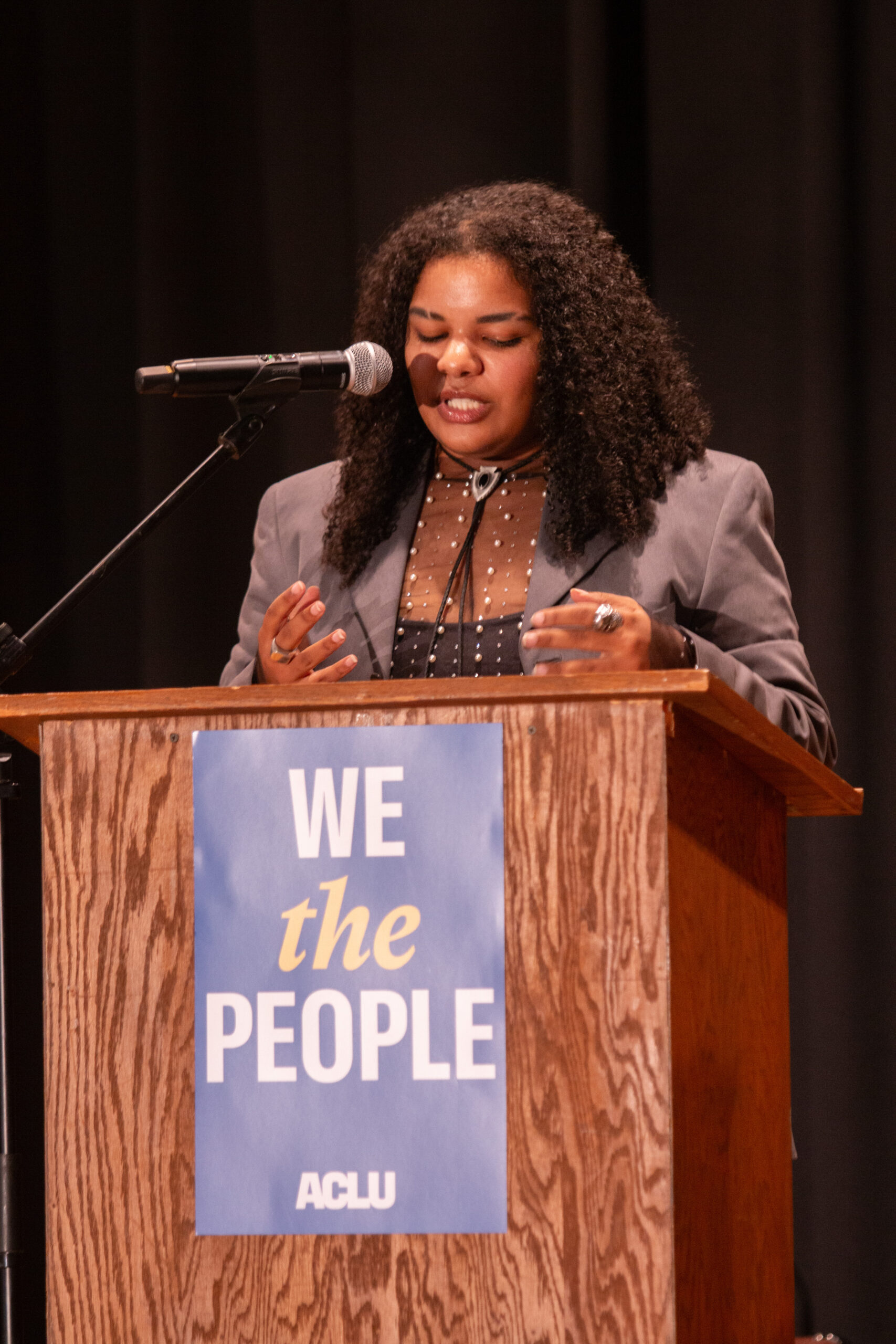 A speaker with long curly black hair wearing a gray blazer over a brown and black polka-dot blouse stands at a wooden podium with a sign reading "WE the PEOPLE" and ACLU logo. They gesture expressively while speaking into a microphone against a dark curtain backdrop.