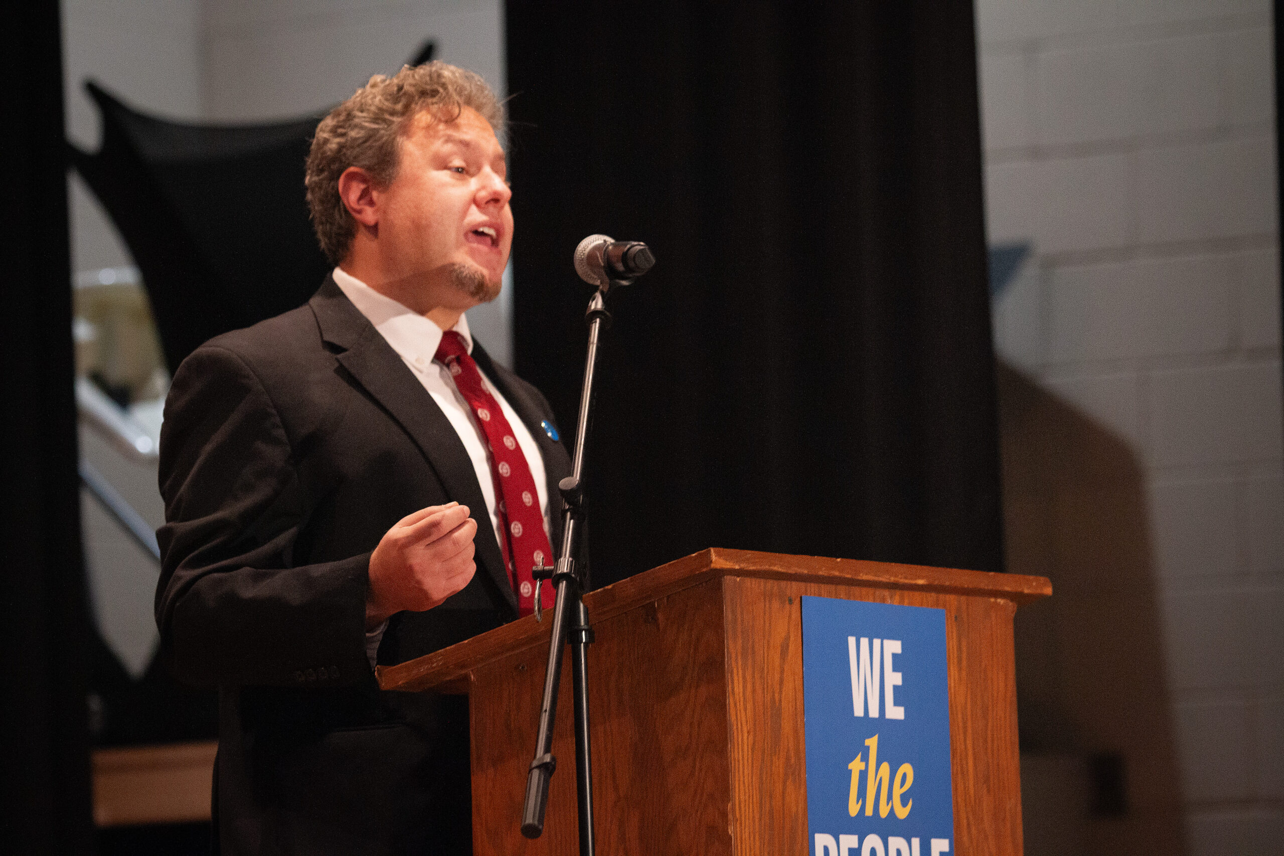 A speaker in a black suit and burgundy patterned tie stands at a wooden podium, gesturing with one hand while speaking passionately into a microphone. Behind them is a sign reading "WE the PEOPLE" with ACLU branding against a dark backdrop.