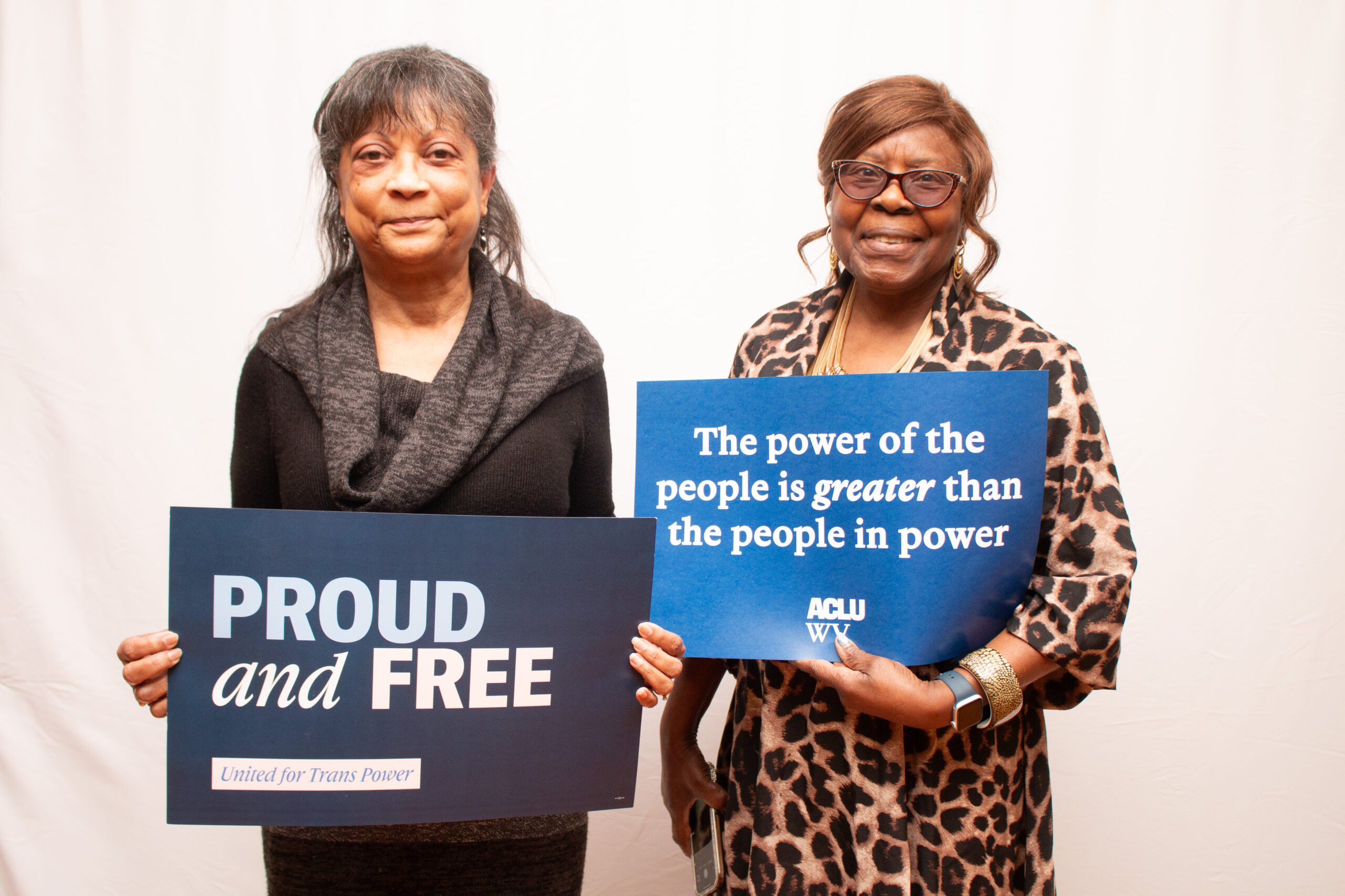 Two people stand against a light backdrop holding advocacy signs. The person on left wears a black top with gray cowl-neck scarf and holds a "PROUD and FREE" sign. The person on right wears a leopard-print blouse with gold jewelry and holds a blue ACLU WV sign reading "The power of the people is greater than the people in power."