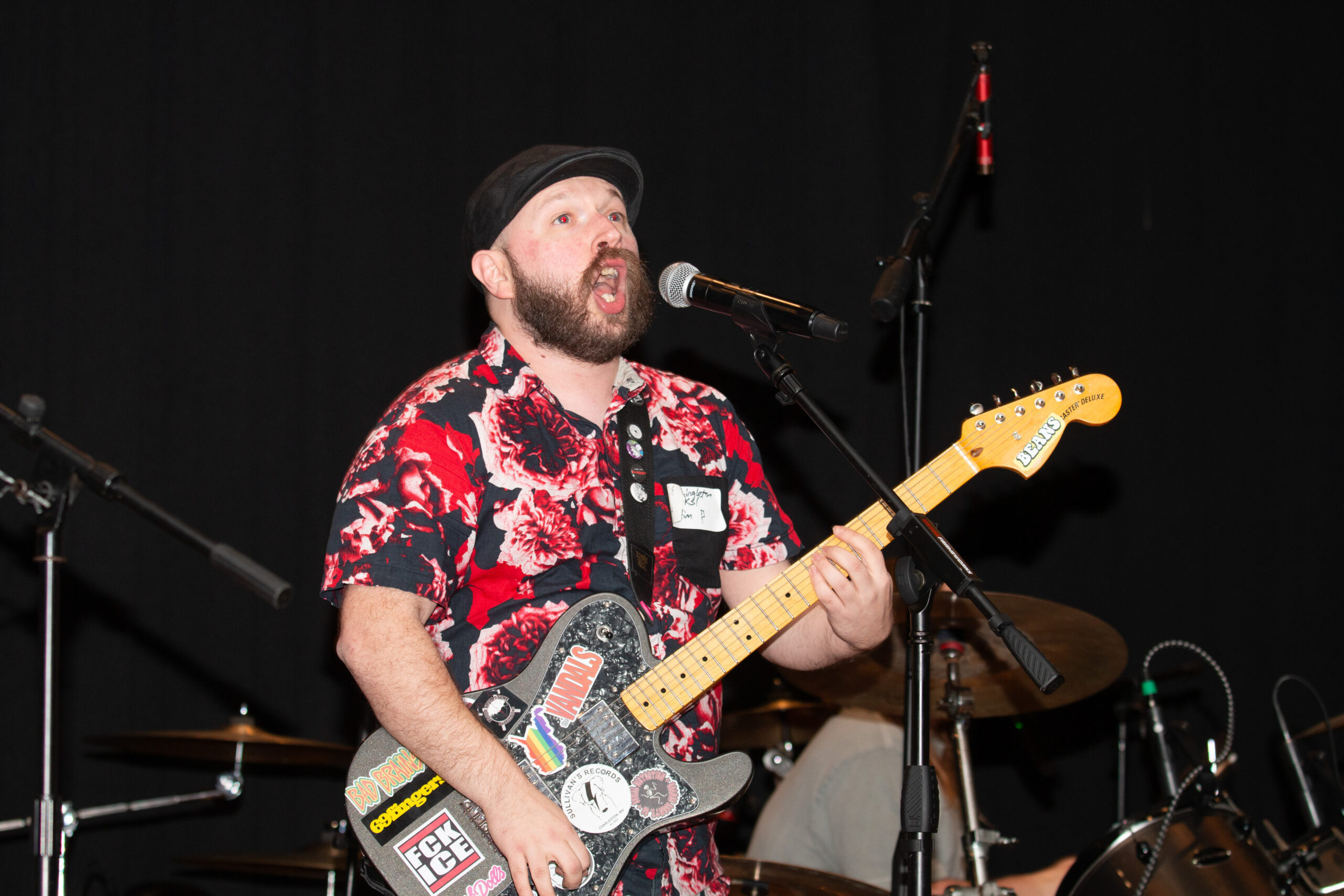 A musician wearing a black beanie and red-and-black floral shirt performs passionately on stage, singing into a microphone while playing a sticker-covered electric guitar. Drum kit and stage equipment are visible in the background against dark curtains.