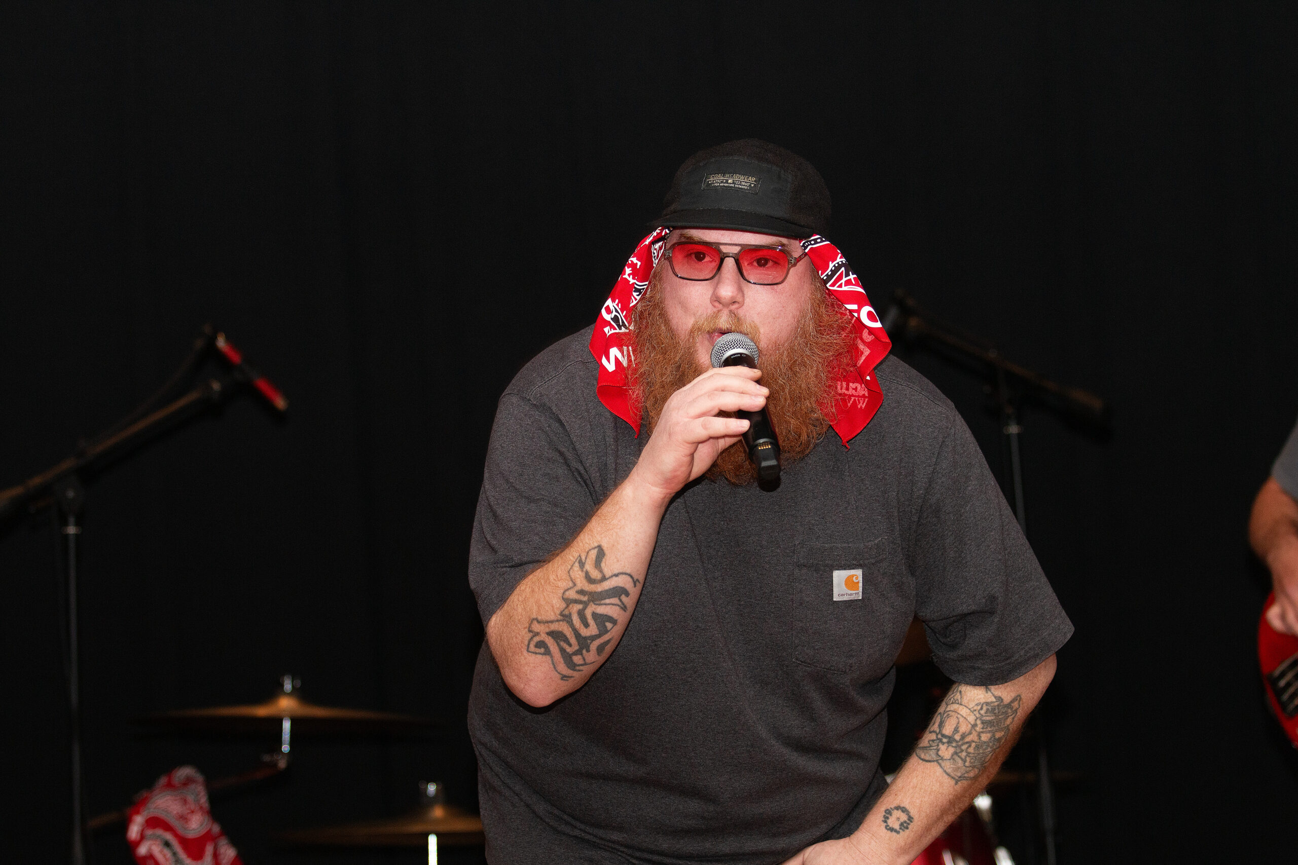 A performer with a full red beard wearing a gray t-shirt, black beanie, and red sunglasses sings into a handheld microphone on stage. Red bandanas hang as decorative elements. They wear visible tattoos on their arms and stand against a dark backdrop.