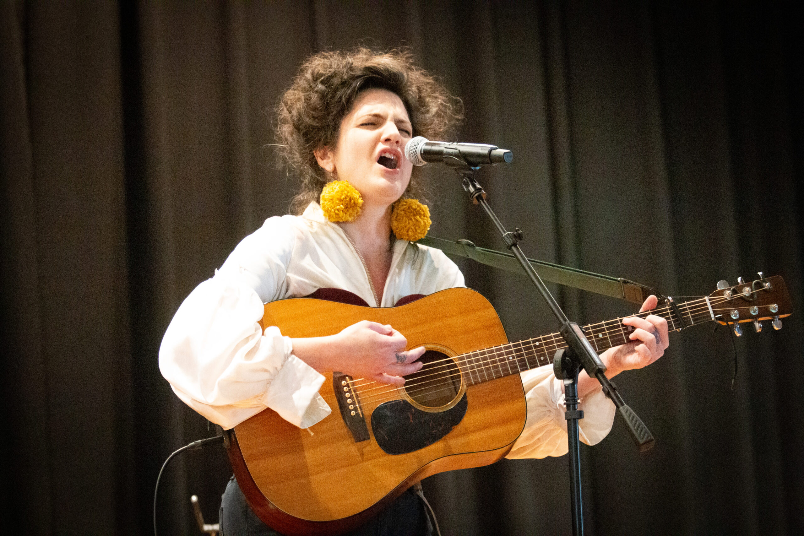A performer with curly dark hair wearing large yellow pom-pom earrings and a white flowing blouse passionately sings into a microphone while playing an acoustic guitar on stage, with a dark curtain backdrop.
