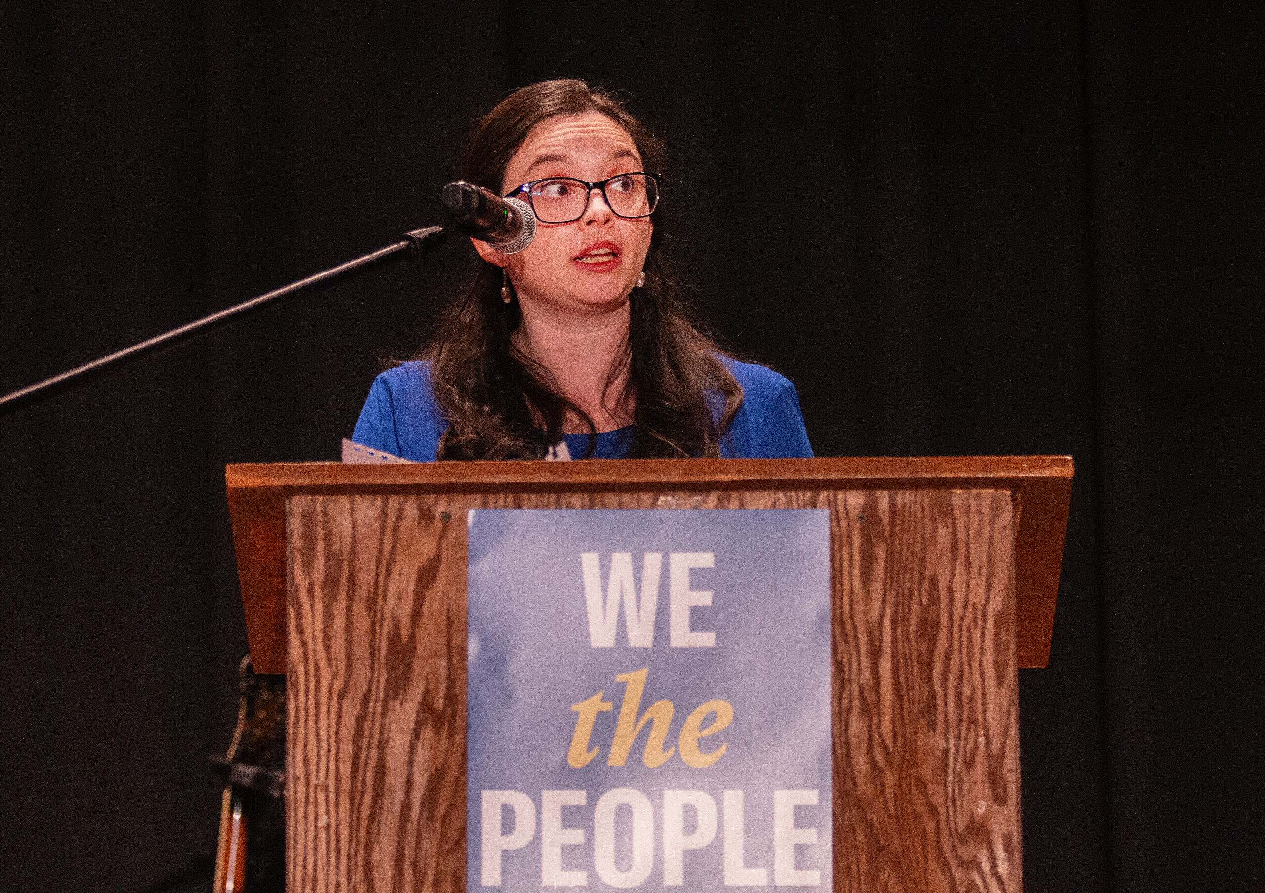 A speaker wearing glasses and a blue dress stands at a wooden podium with a microphone, looking upward thoughtfully while speaking. The podium displays a sign reading "WE the PEOPLE" in blue, yellow, and white text with ACLU branding.
