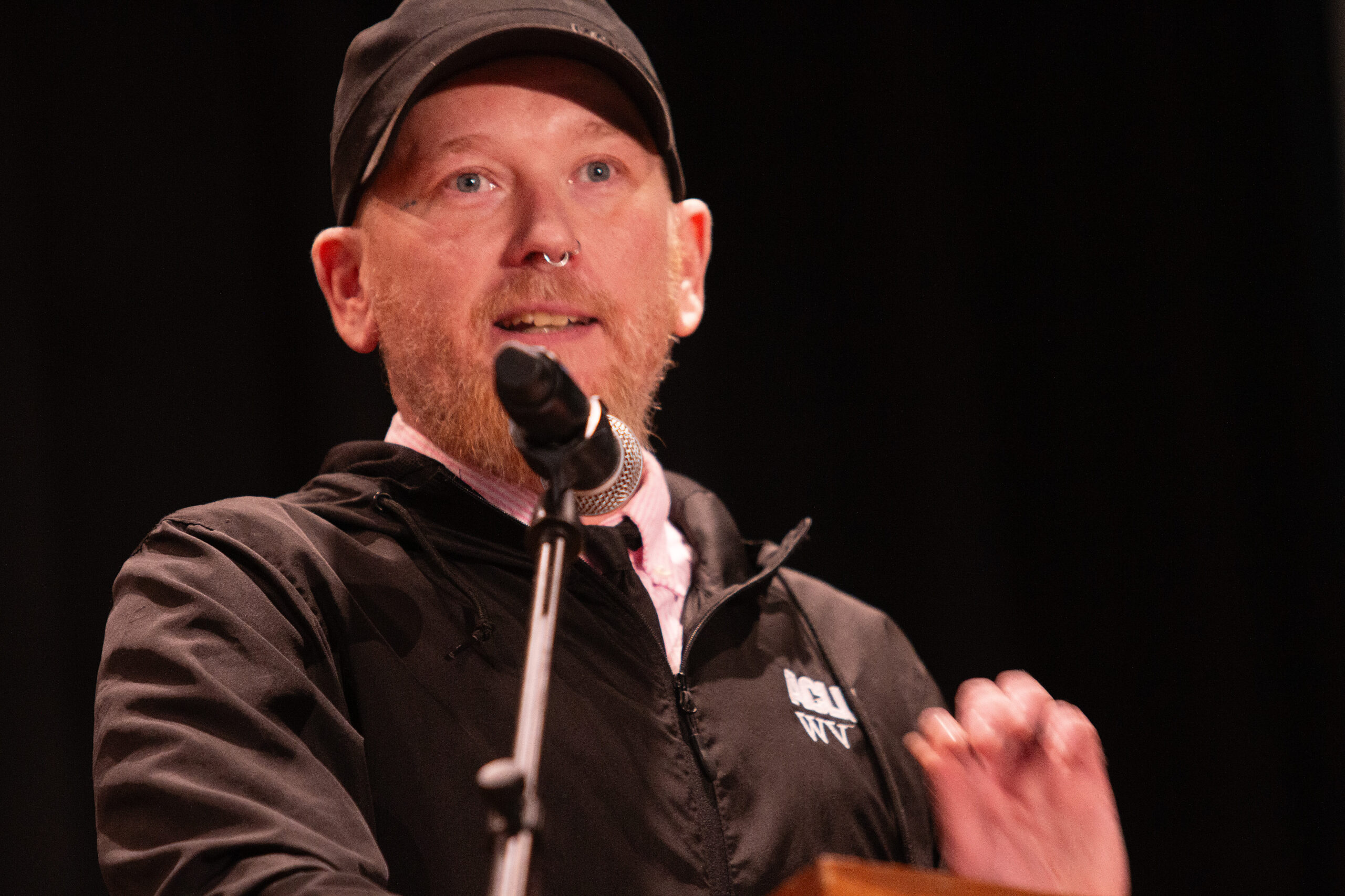 A speaker wearing a brown baseball cap, brown jacket with "ACLU WV" text, and nose ring speaks into a microphone on stage with one hand raised in a gesturing motion. Black backdrop behind them.