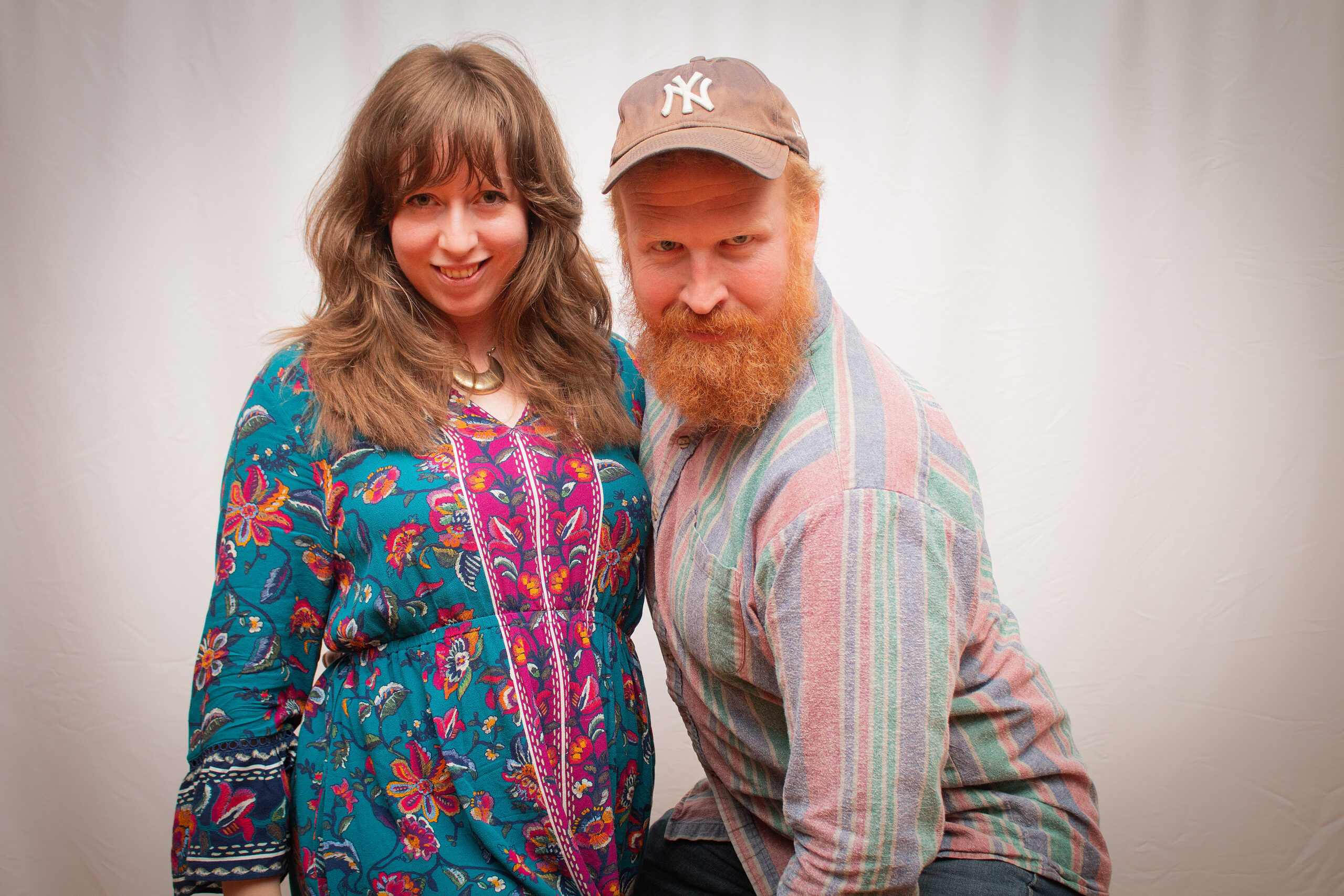 Two people pose together against a light backdrop. The person on left wears a vibrant turquoise and pink floral-patterned dress with embroidered details and a crescent moon necklace. The person on right wears a brown cap and multicolored striped shirt, sporting a full red beard.