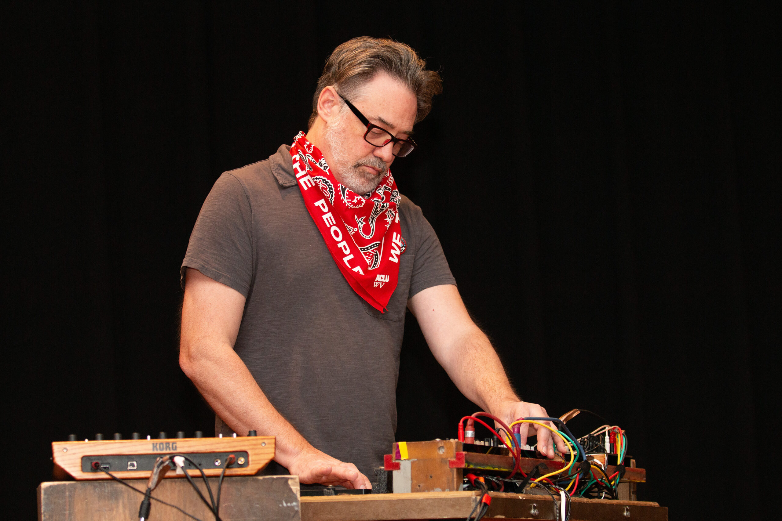 A person wearing glasses and a gray t-shirt with a red "WE THE PEOPLE" bandana tied around their neck operates electronic music equipment on a table. Multiple colorful wires connect various synthesizer modules mounted on wooden boards against a black backdrop.