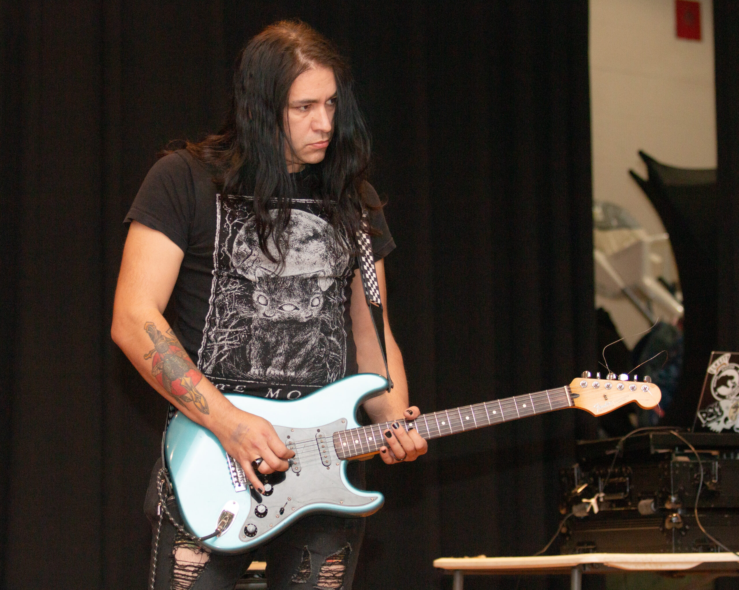 A musician with long dark hair wearing a black graphic t-shirt plays a light blue electric guitar with white pickguard on stage. Ripped black pants visible, performing against dark curtains with stage equipment in the background.