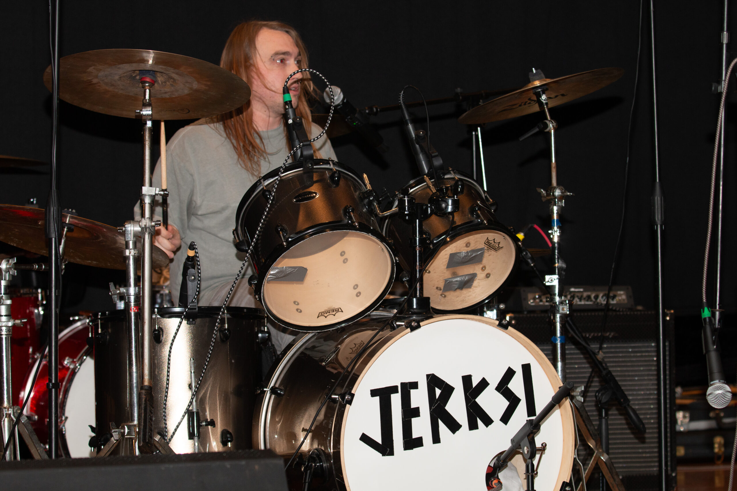 A drummer with shoulder-length hair wearing a gray t-shirt sits behind a drum kit decorated with chains. The bass drum displays "JERRU" in bold black letters. Multiple cymbals and microphones are positioned around the kit against a black backdrop.