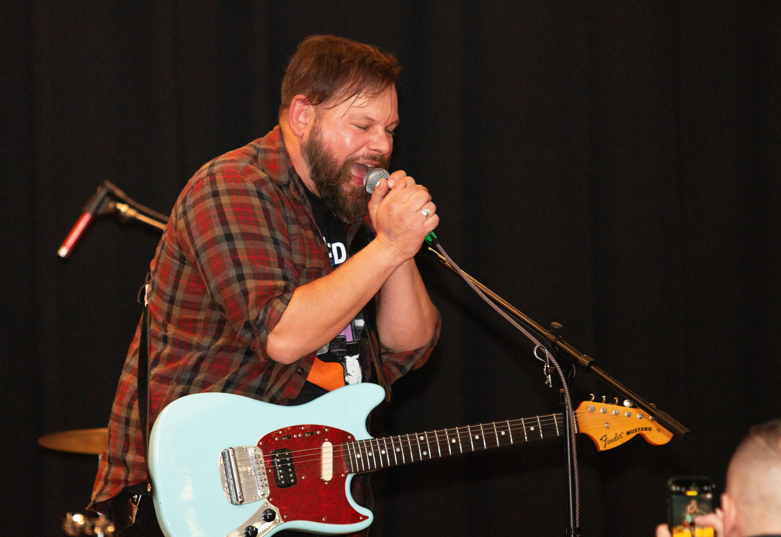 A musician wearing a plaid flannel shirt over a graphic t-shirt sings intensely into a microphone while playing a light blue electric guitar with red pickguard. Stage lighting creates warm tones against the dark performance space.