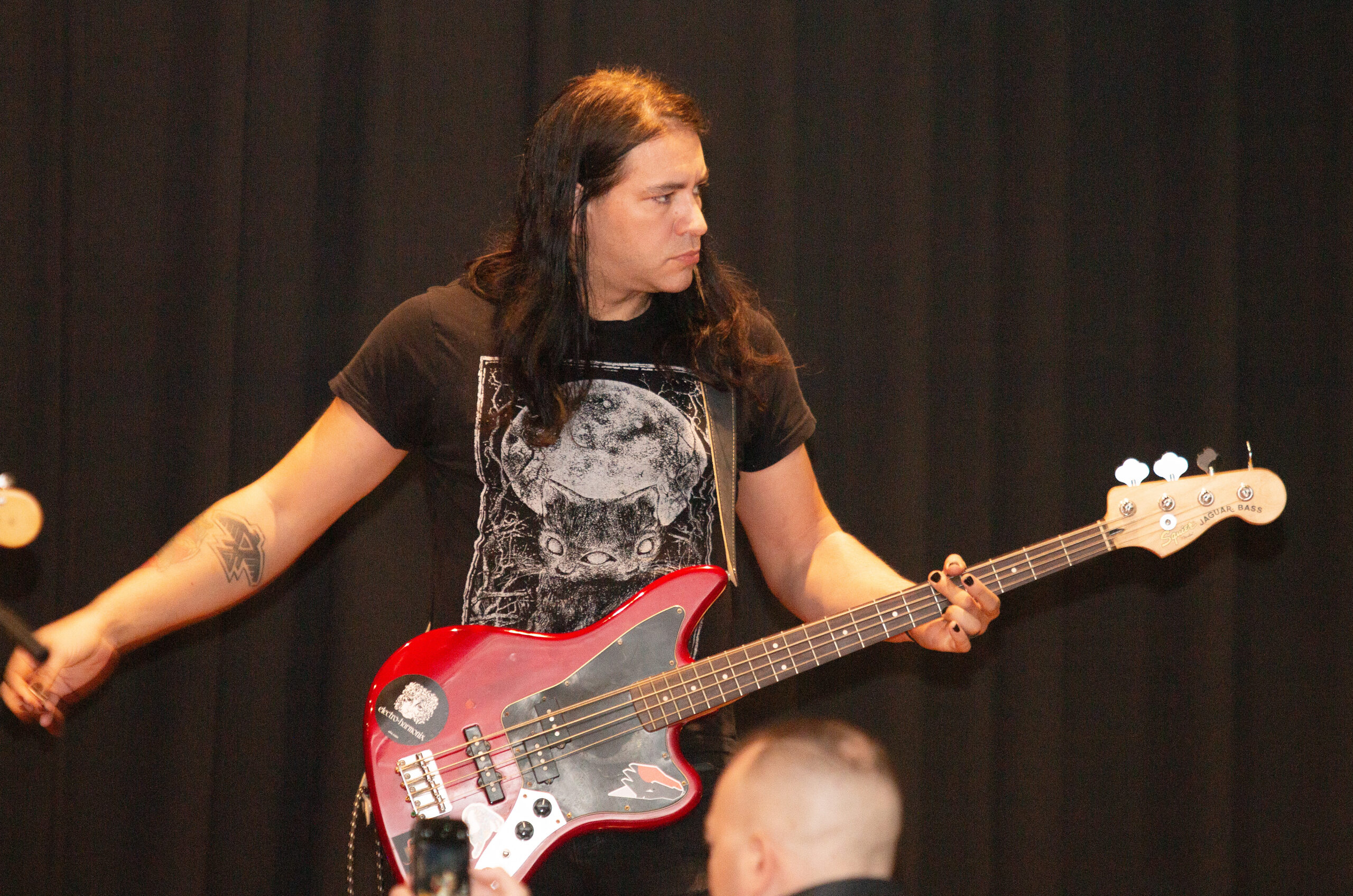 A musician with long dark hair wearing a black graphic t-shirt plays a red-to-pink gradient bass guitar on stage. They wear a chain on their hip and black pants, performing against dark curtains with stage equipment visible in the background.