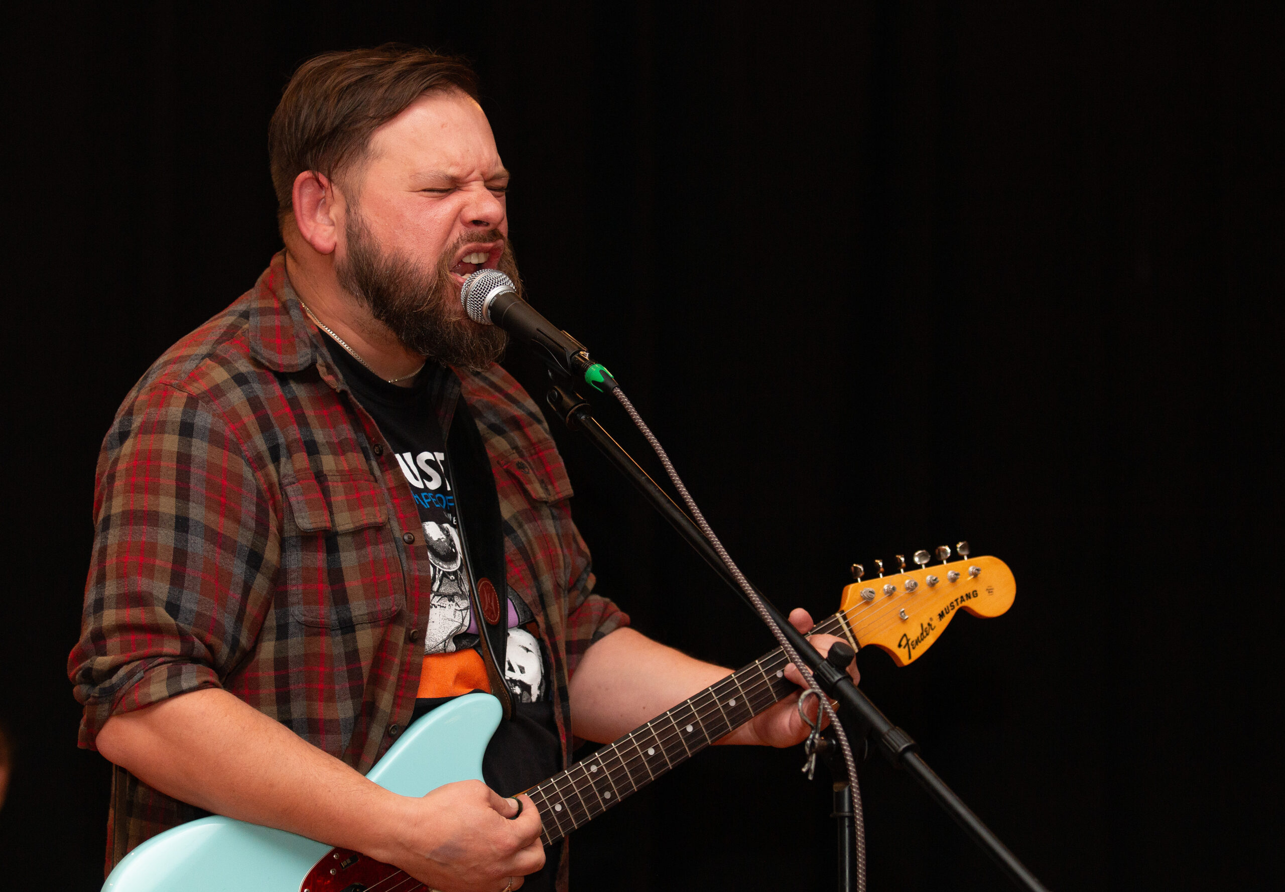 A vocalist wearing a plaid flannel shirt sings passionately into a microphone while playing a light blue electric guitar. Eyes closed in concentration, they wear a graphic t-shirt underneath, performing on stage with a black backdrop.
