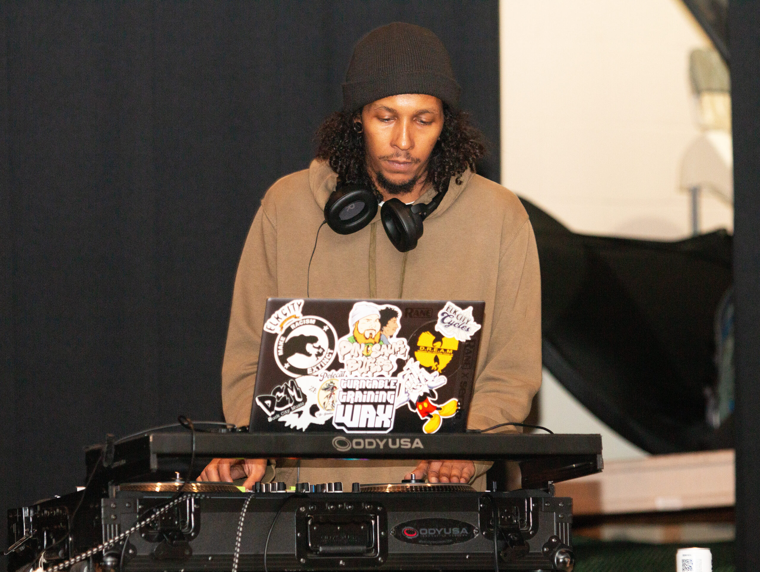 A DJ wearing a brown beanie and tan hoodie with headphones around their neck operates turntables and mixing equipment. The laptop in front of them is covered in various colorful stickers including cartoon characters. They focus on their equipment with eyes downcast.