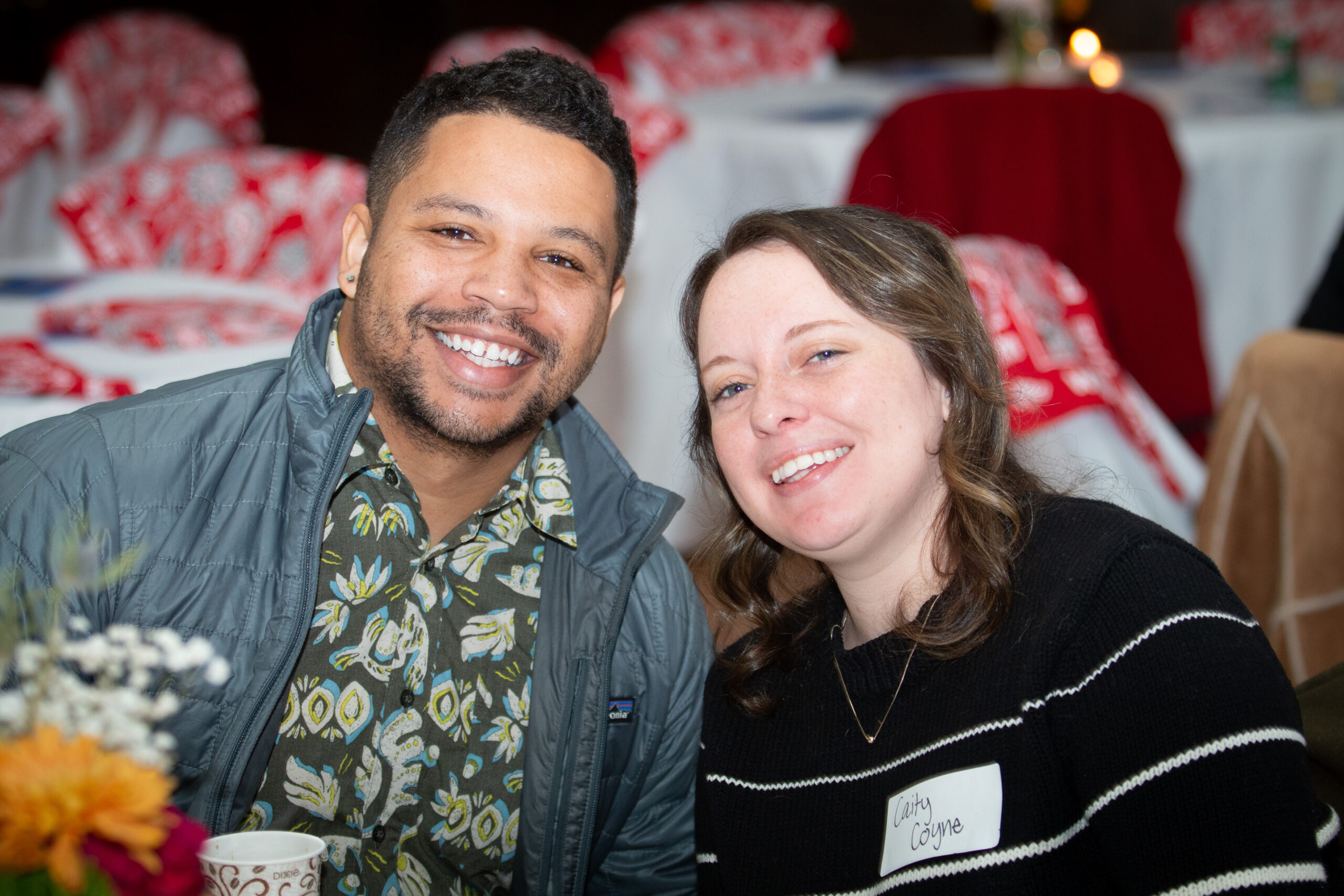 Two people smile warmly at the camera at what appears to be a festive gathering. The person on left wears a blue vest over a floral shirt, while the person on right wears a black striped sweater with a name tag. Red and white decorations are visible in the blurred background.