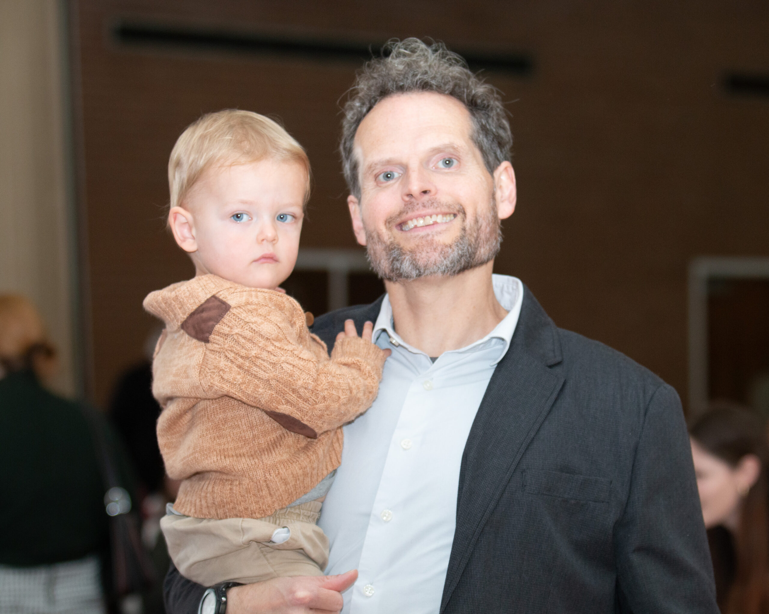 An adult with curly gray hair and beard wearing a dark blazer over a light blue shirt holds a young child with blonde hair in their arms. The child wears a tan knitted sweater and khaki pants. They stand in an indoor venue with blurred event attendees visible in the background.