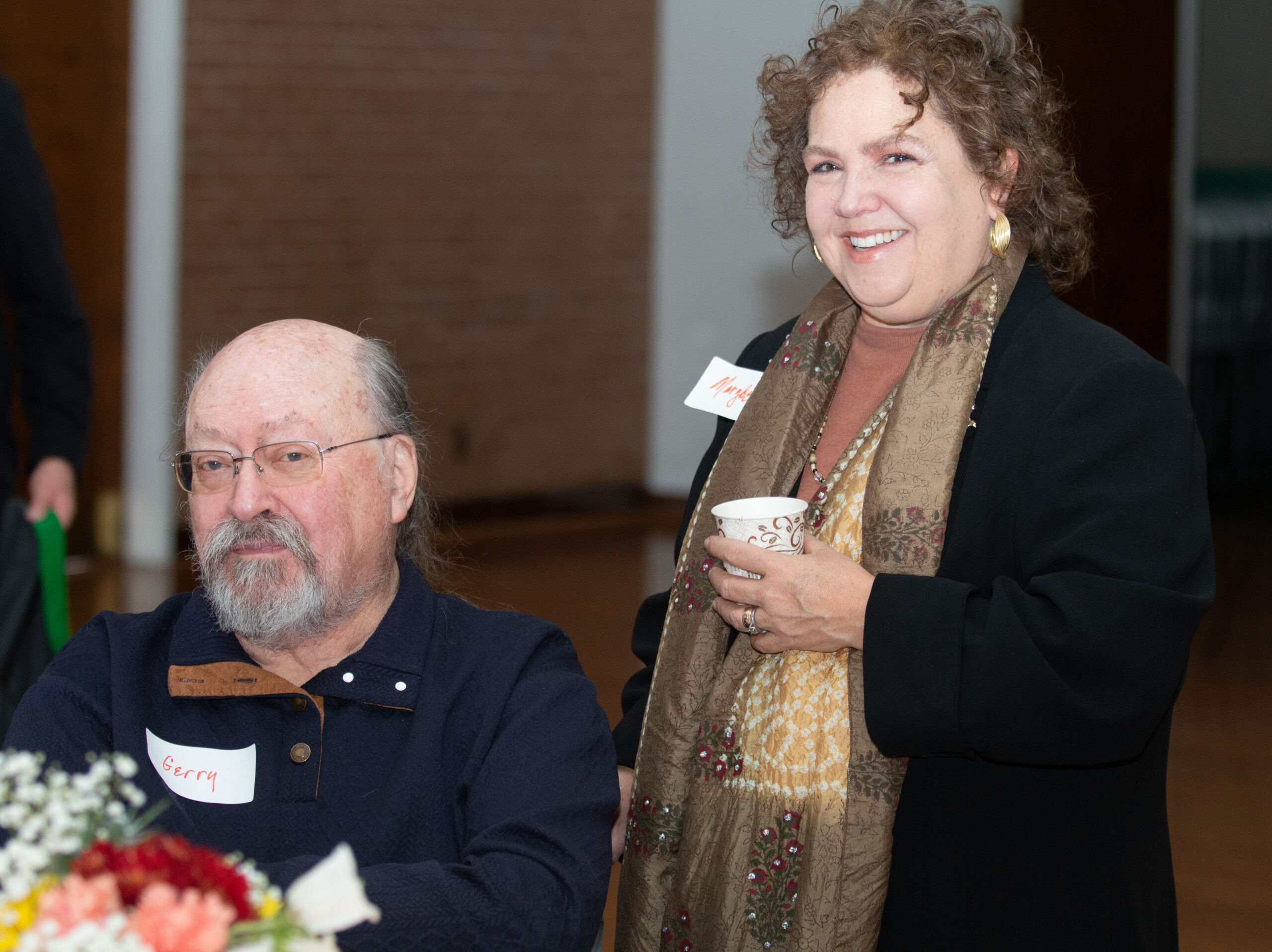 Two people stand together at an indoor event. The person on left, seated in a wheelchair, wears glasses and a dark polo with "Berry" name tag. The person on right with curly hair wears gold hoop earrings, a black jacket over an embellished scarf, and yellow patterned skirt while holding a coffee cup.