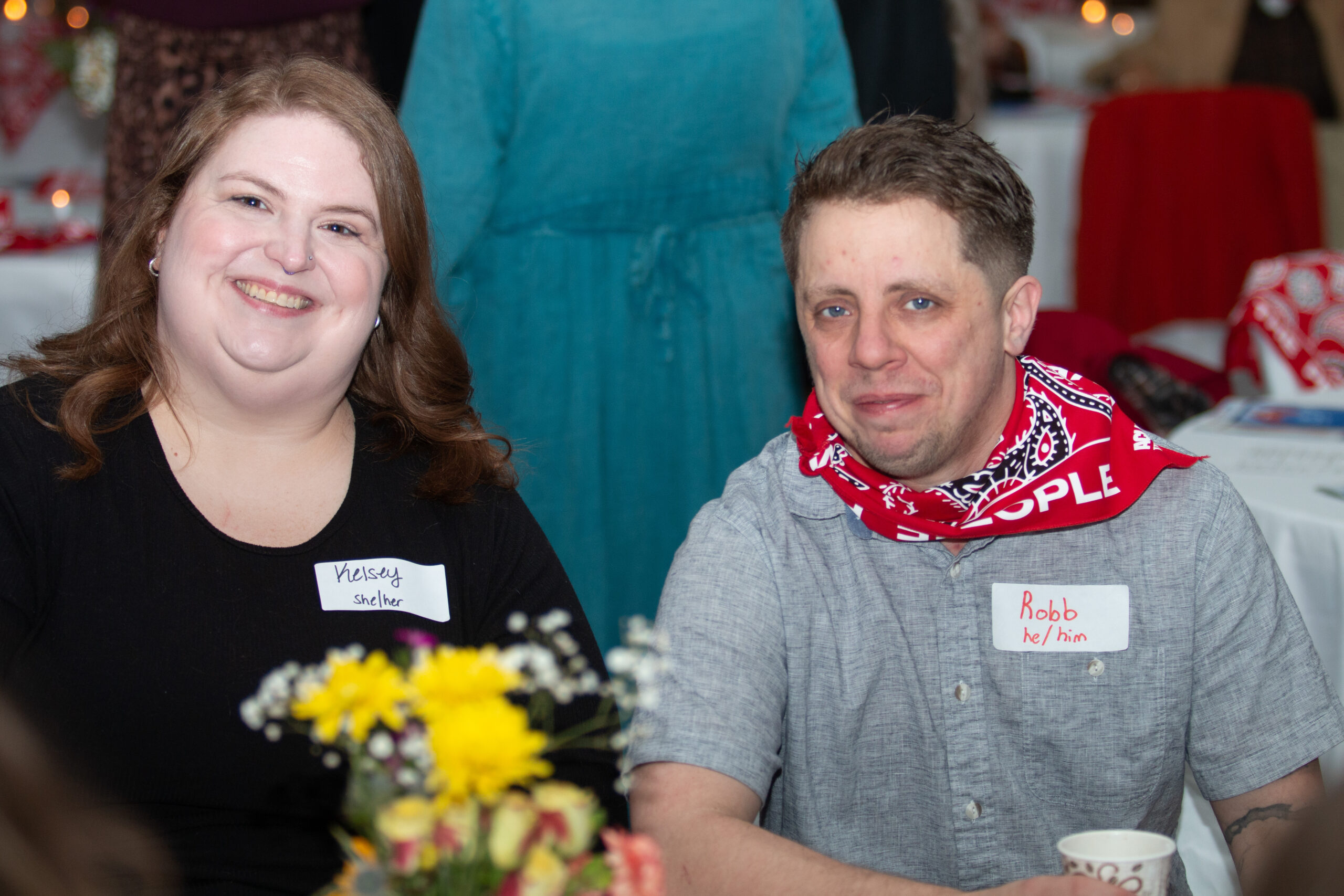 Two people sit at a table with floral centerpieces at an event. The person on left wearing black with "Kelsey shelter" name tag smiles broadly. The person on right wearing a gray shirt with red "WE THE PEOPLE" bandana around neck has a name tag reading "Robb he/him." Event decorations visible in background.