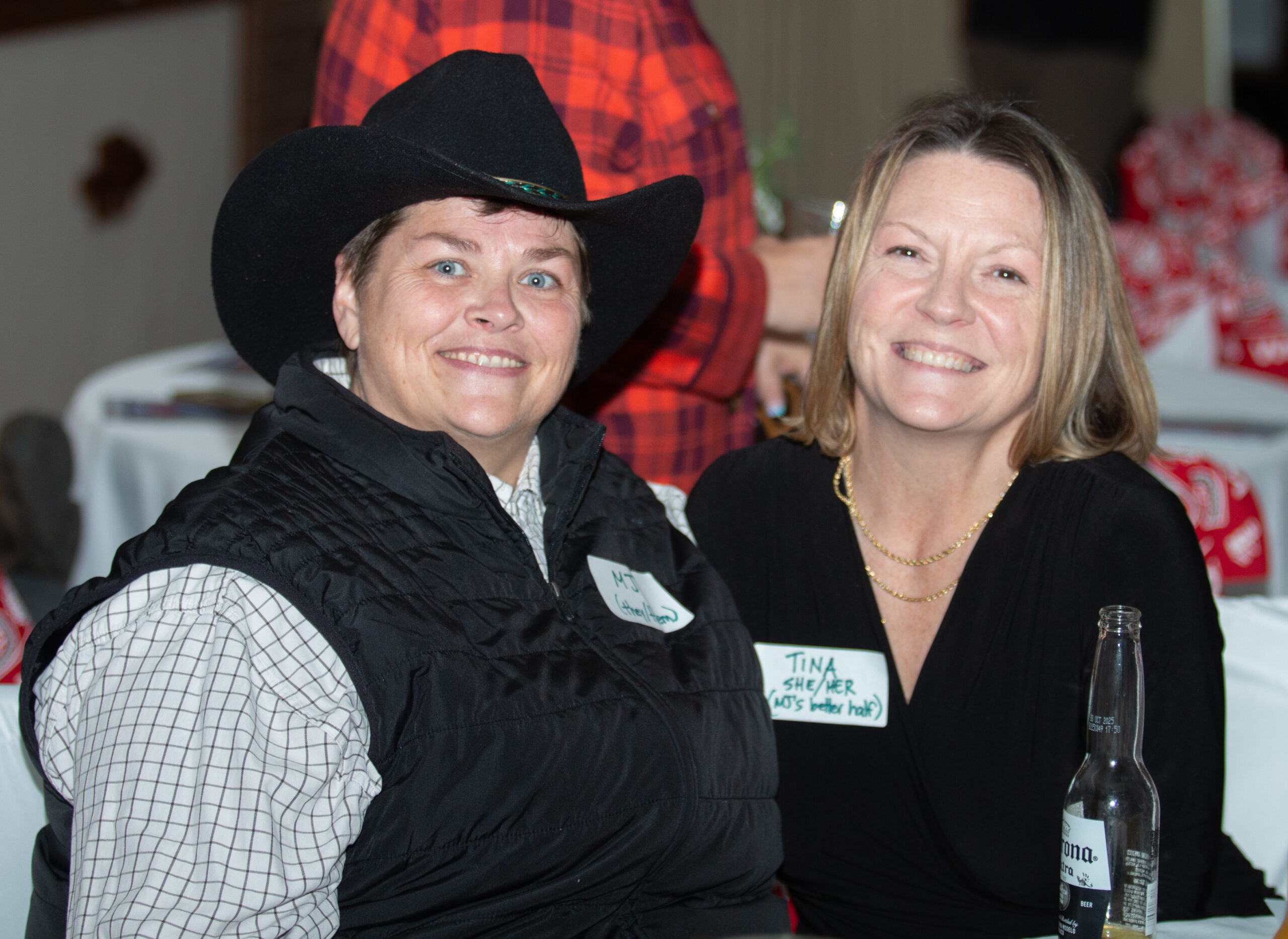 Two people smile at camera in an event space. The person on left wears a black cowboy hat, black quilted vest, and plaid shirt underneath, with a name tag. The person on right wears a black top with gold chain necklace and holds a beer bottle. Red and white decorations visible in background.