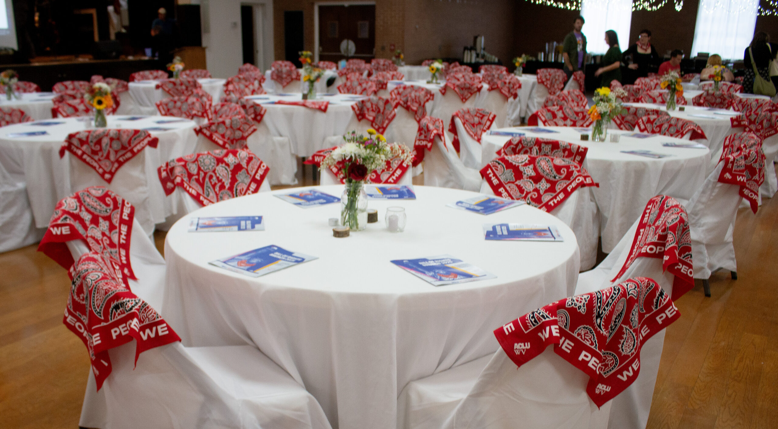 Wide view of event space with numerous round tables covered in white tablecloths. Each table features red "WE THE PEOPLE" ACLU WV bandanas draped on chairs, small floral centerpieces with sunflowers, and blue programs. String lights hang overhead. Attendees visible mingling in the background under exposed ceiling with industrial elements.