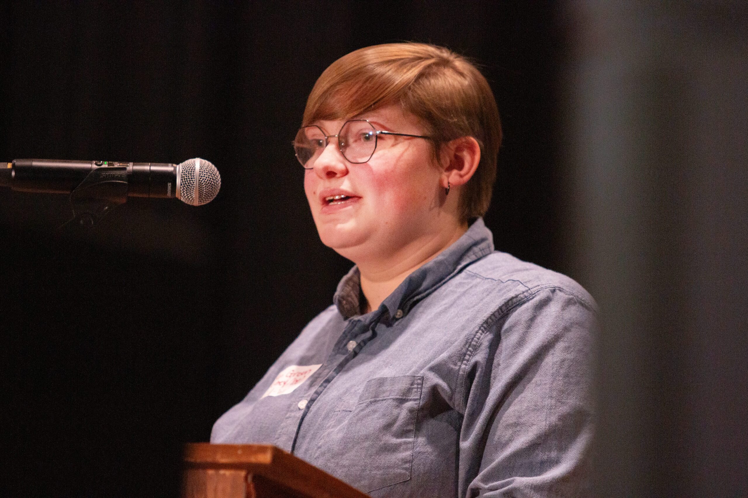 A person with short hair and glasses wearing a light blue chambray shirt speaks or sings at a wooden podium on stage. A microphone on stand is positioned nearby. Dark curtain backdrop behind them creates a simple, focused composition.