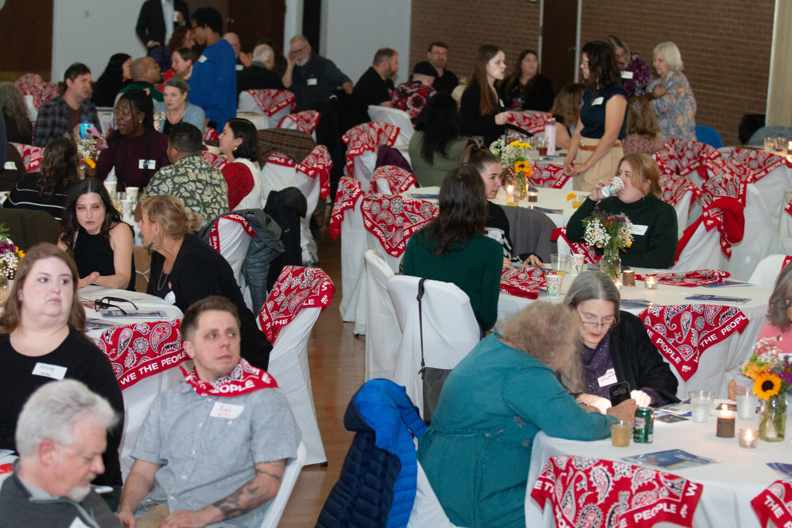 Crowded event hall with attendees seated at round tables covered with white tablecloths and red "WE THE PEOPLE" bandanas on chairs. Small floral centerpieces with sunflowers and candles decorate each table. Diverse group mingles and converses, with some standing and others seated. Exposed brick wall visible in background.