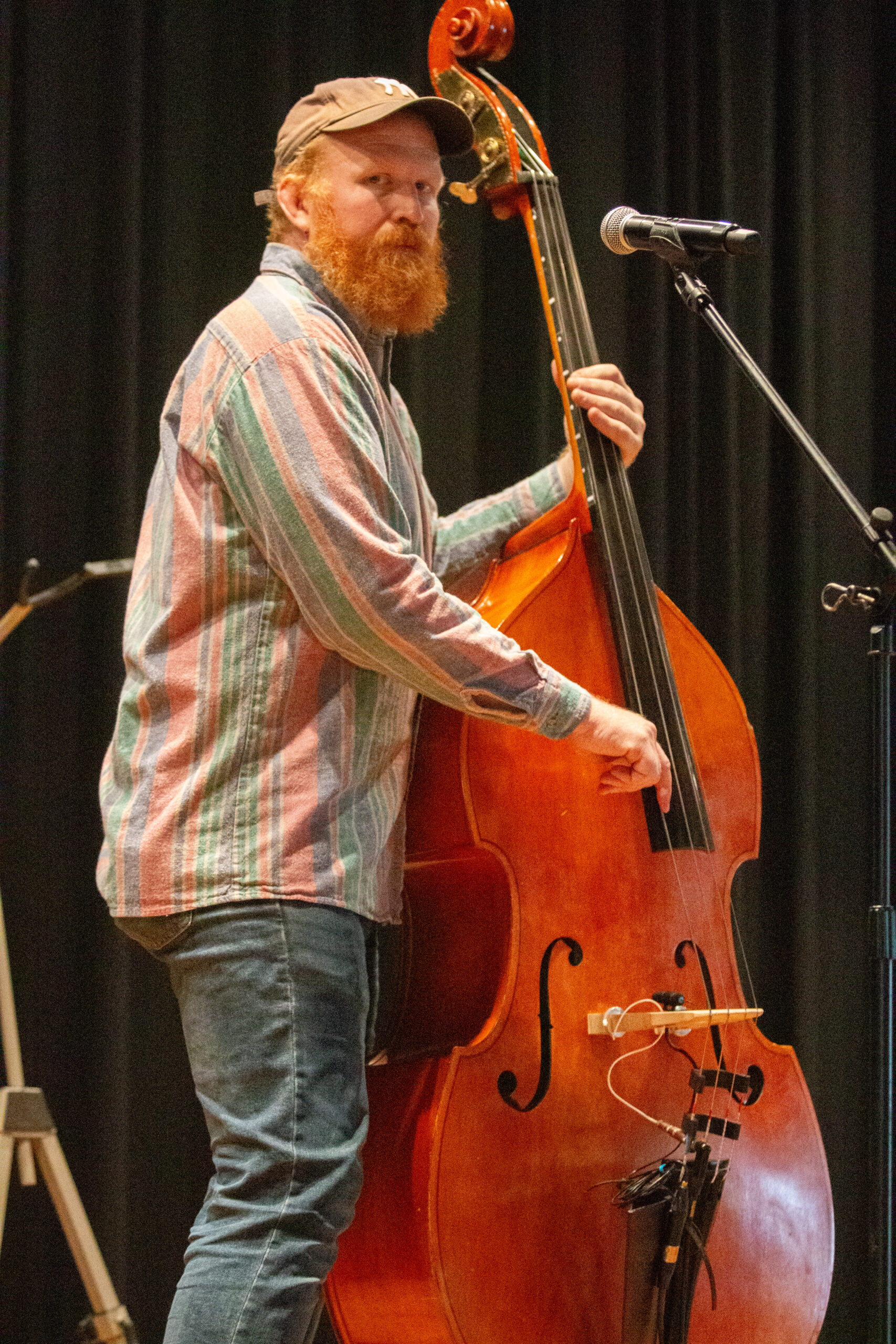 A musician with red beard wearing a tan baseball cap and pastel striped button-up shirt plays upright bass on stage. A microphone stands nearby. They perform against a black curtain backdrop, focused on their instrument with stage lighting creating warm tones.
