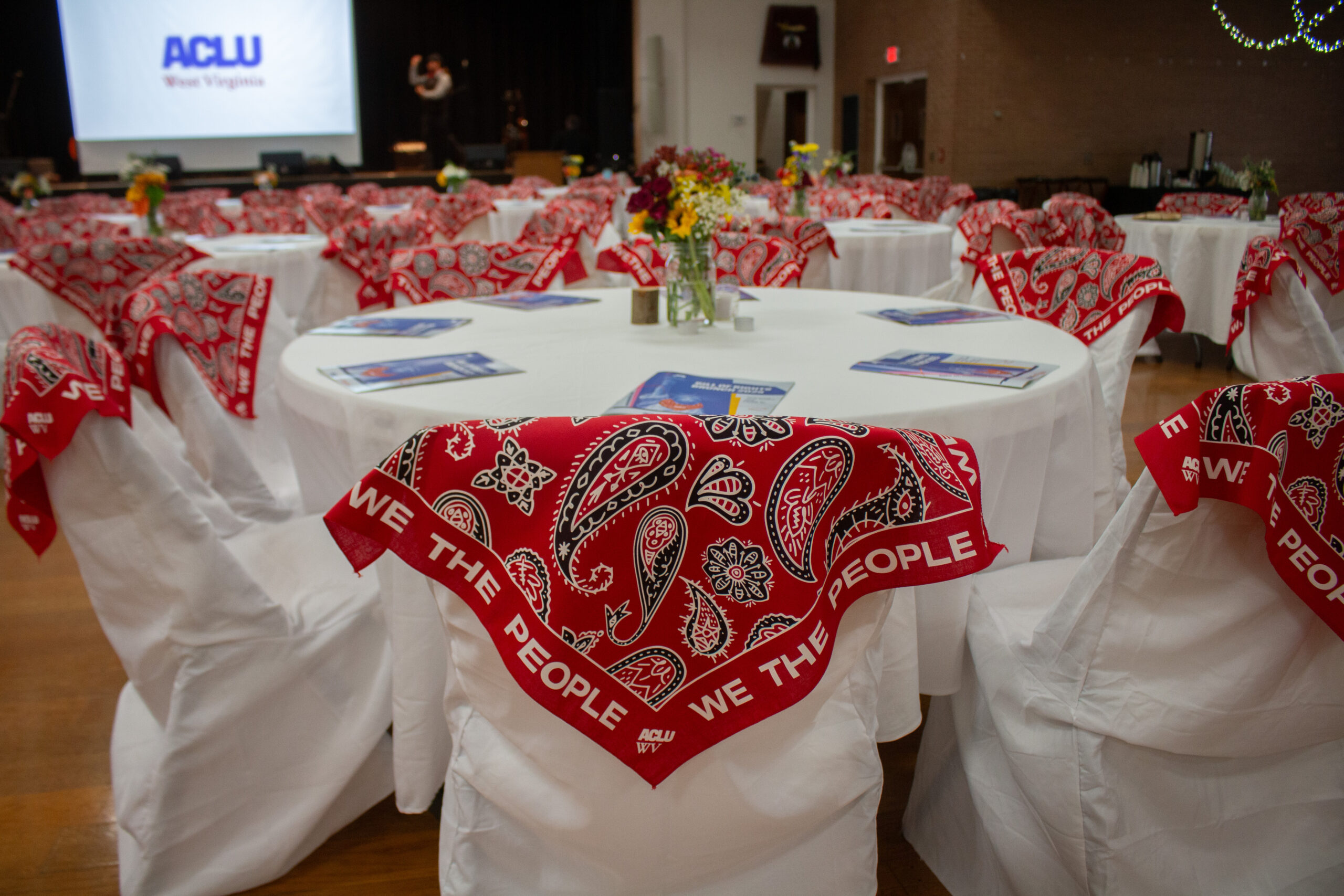 Close-up detail shot of event seating with white folding chairs decorated with red paisley bandanas reading "WE THE PEOPLE" with ACLU WV branding. Round tables covered with white tablecloths feature blue programs, small floral centerpieces with colorful blooms, and candles. ACLU logo visible on projection screen in blurred background.