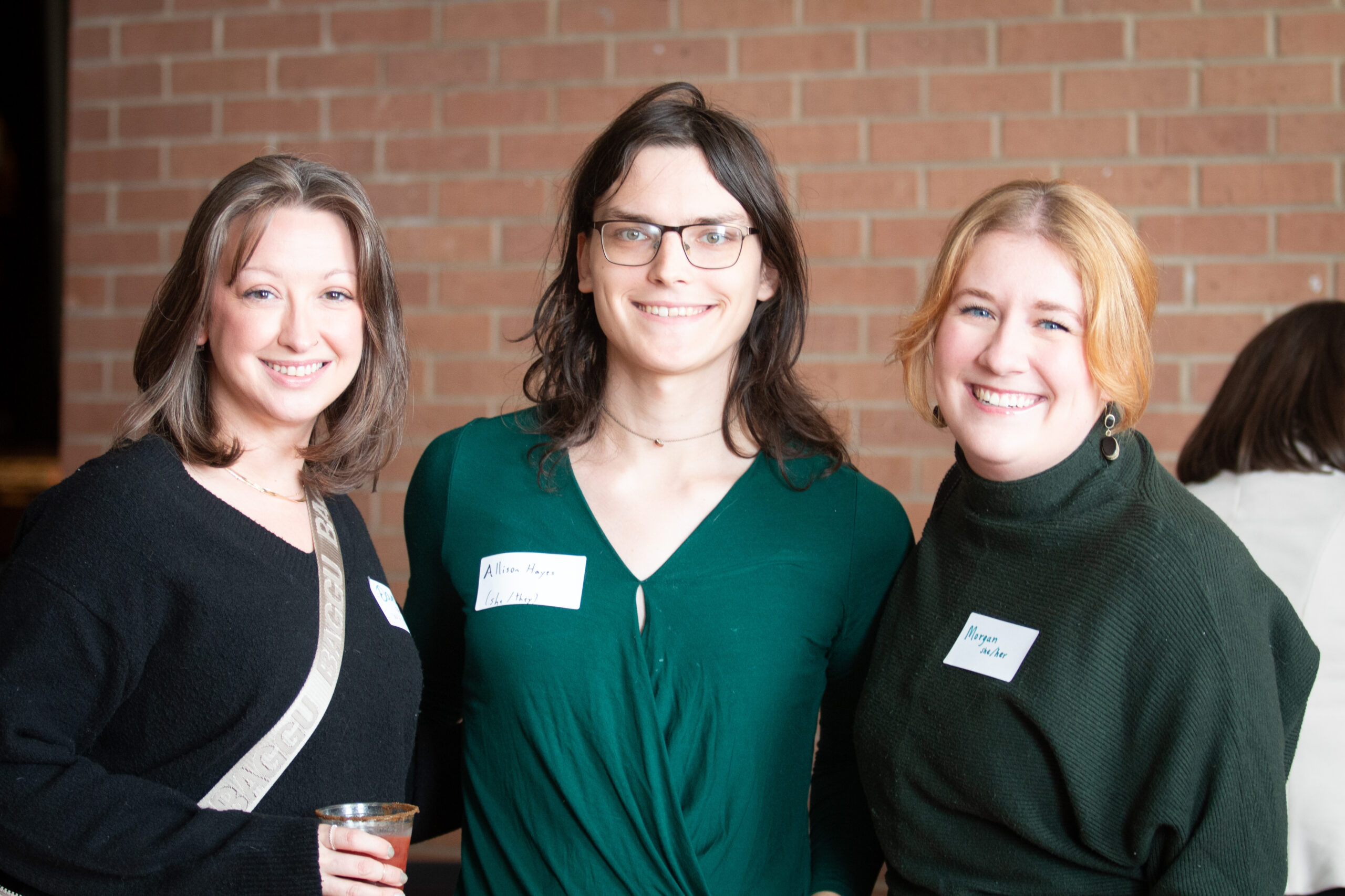 Three people stand together smiling in front of an exposed brick wall, all wearing name tags. From left to right: person in black top with cross-body bag, person in center wearing green v-neck dress and glasses, person on right in dark green turtleneck sweater.