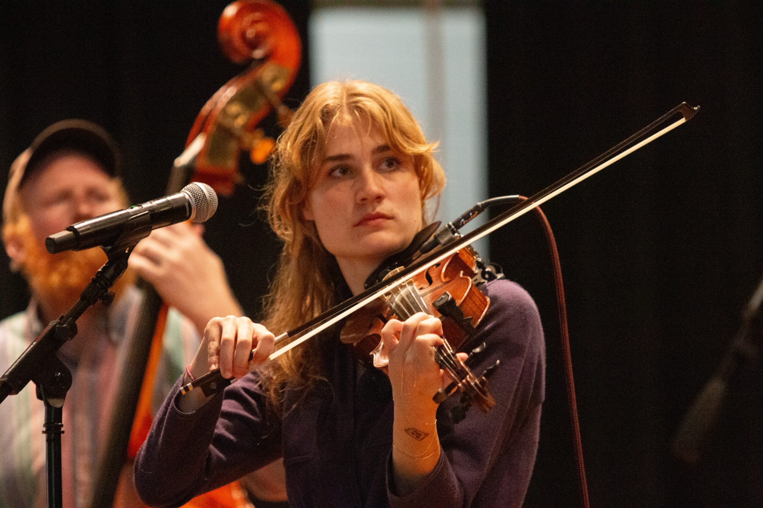 A musician with wavy reddish-blonde hair wearing a purple long-sleeve top performs on violin, bow raised mid-stroke. They stand at a microphone with focused, upward gaze. An upright bass player visible but blurred in the background against dark stage setting.