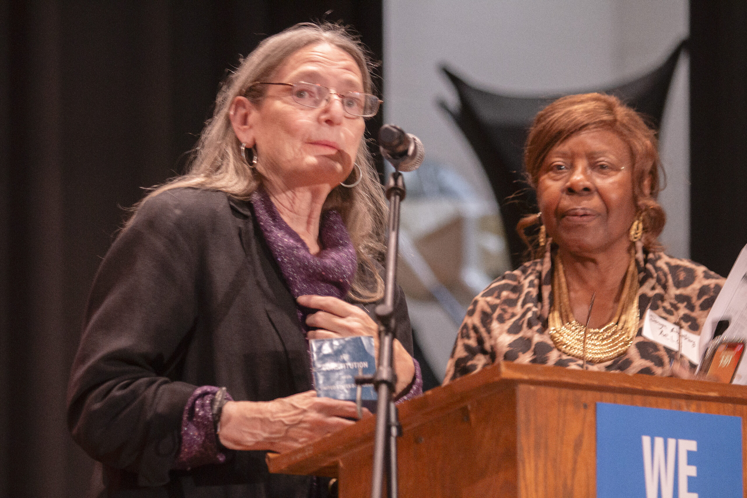 Two people stand at a wooden podium on stage. The person at the podium with long gray hair and glasses wears a black jacket over purple sparkly top, holding what appears to be a Constitution booklet. Another person in leopard print with gold jewelry stands beside them, both at a microphone.