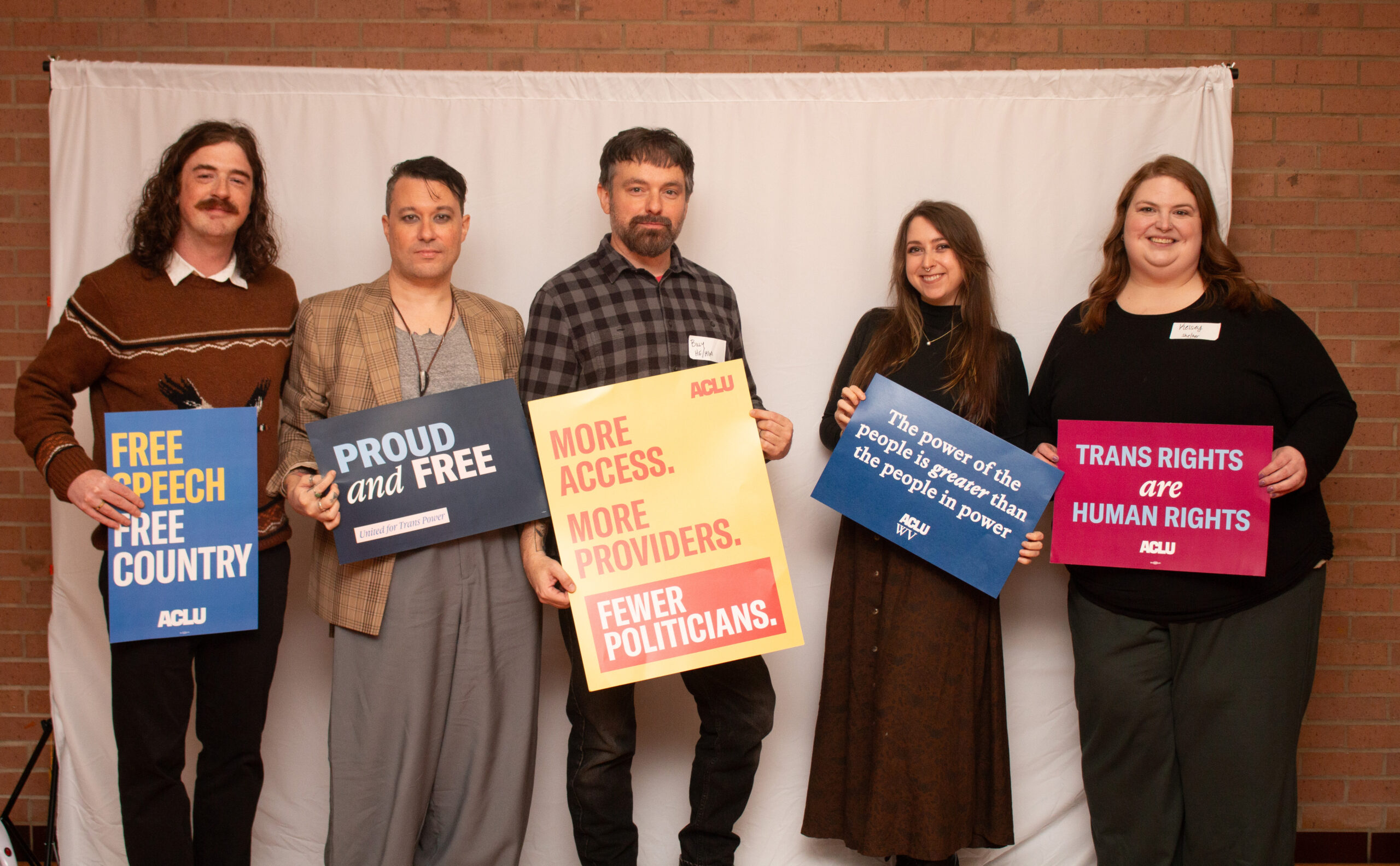 Five people stand in a line against a white backdrop with exposed brick wall visible at left, holding various ACLU advocacy signs. From left: person in brown sweater with "FREE SPEECH FREE COUNTRY," person in tan blazer with "PROUD and FREE," person in center with plaid shirt holding yellow "MORE ACCESS. MORE PROVIDERS. FEWER POLITICIANS.," person in black holding blue "The power of the people is greater than the people in power" sign, and person on right in black holding pink "TRANS RIGHTS are HUMAN RIGHTS" sign.