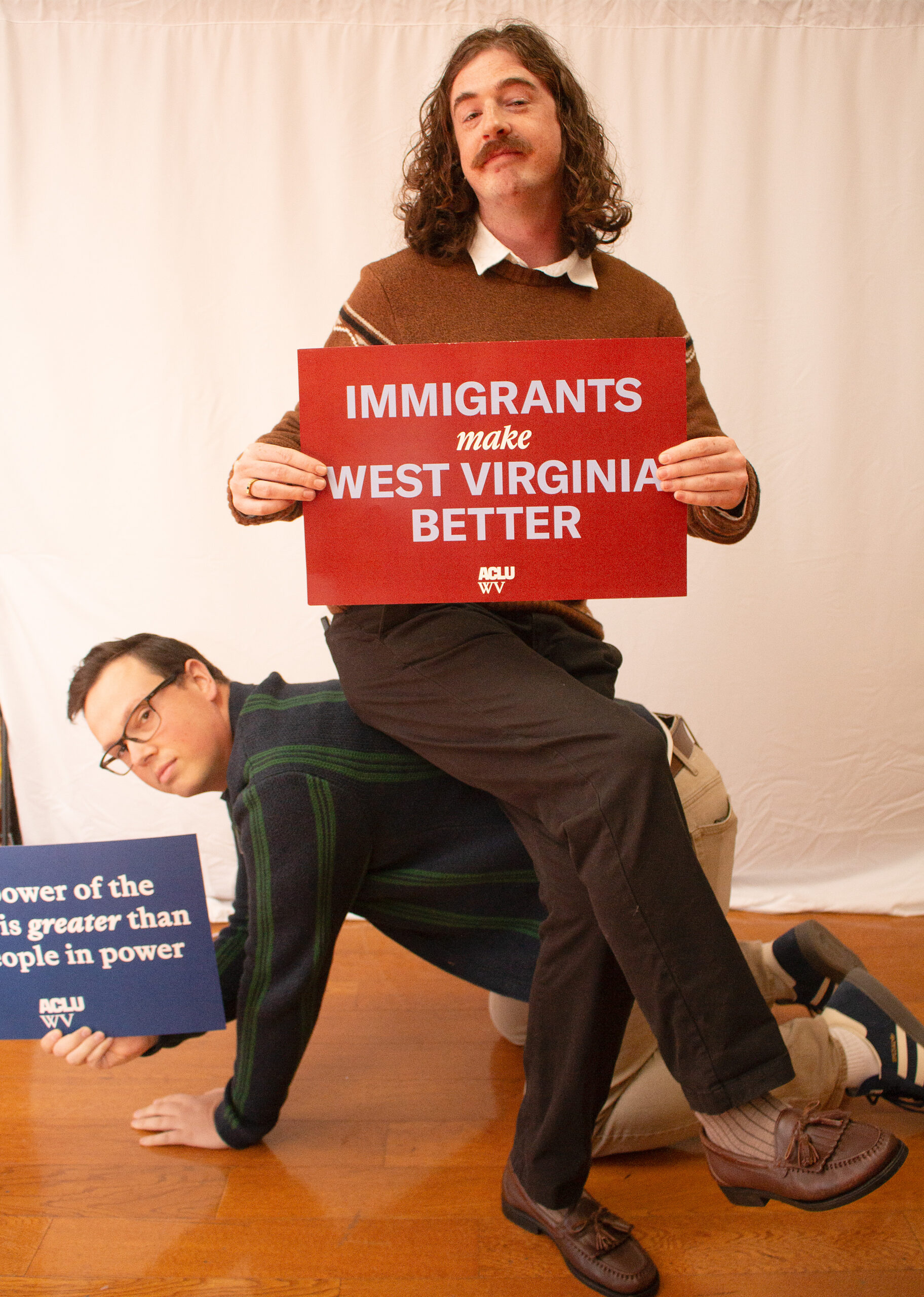 Playful photo of two people in front of white backdrop. Person in foreground sitting on their companion's back wears brown sweater with white collar and mustache, holding red ACLU WV sign reading "IMMIGRANTS make WEST VIRGINIA BETTER." Person below wears green and black striped sweater, supporting them while holding a partially visible blue sign.