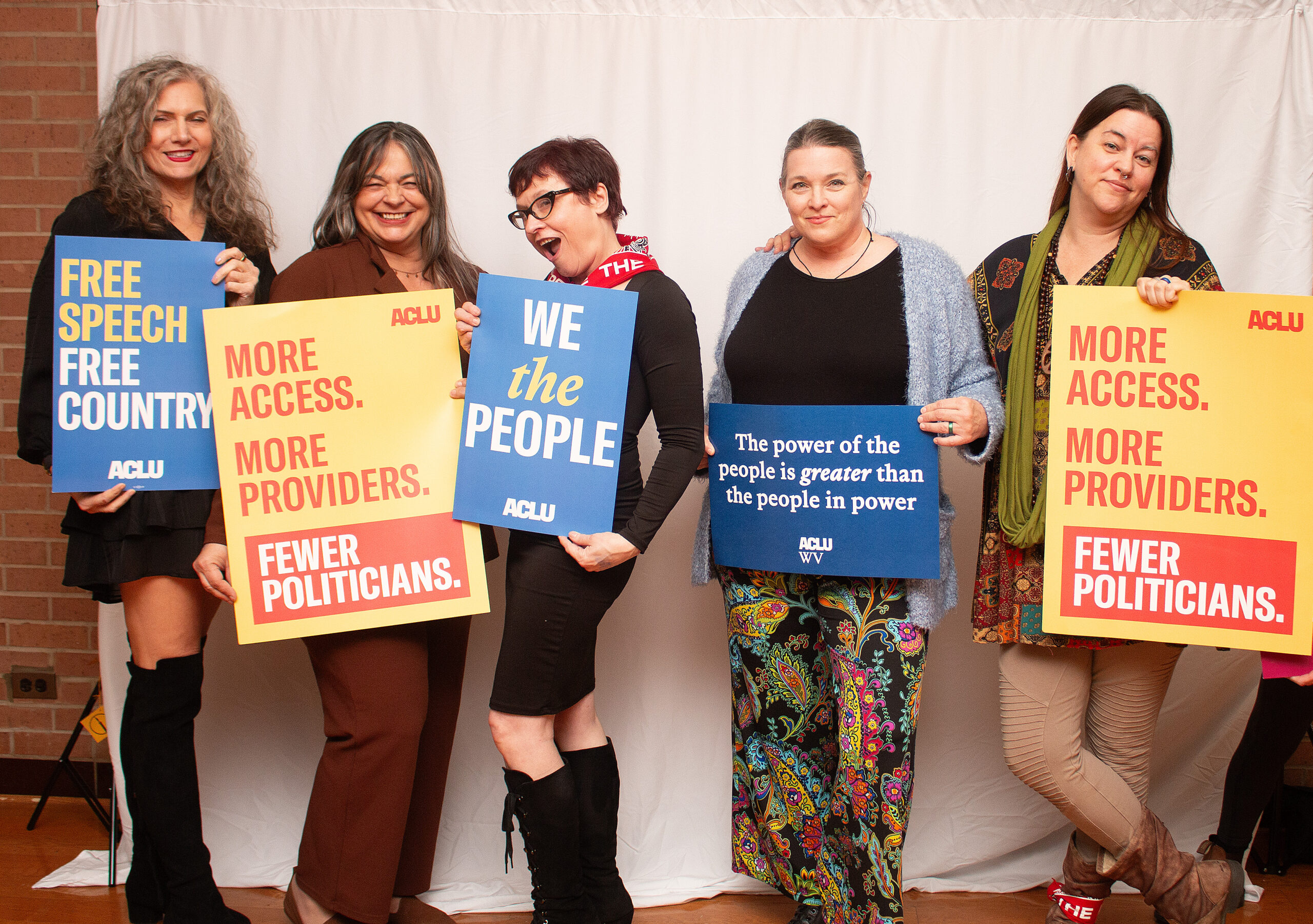 Five people pose together holding various ACLU advocacy signs against a white backdrop with exposed brick. From left to right they hold signs reading "FREE SPEECH FREE COUNTRY," yellow "MORE ACCESS. MORE PROVIDERS. FEWER POLITICIANS.," blue "WE the PEOPLE," blue "The power of the people is greater than the people in power," and yellow "MORE ACCESS. MORE PROVIDERS. FEWER POLITICIANS." Group displays mix of formal and casual attire.