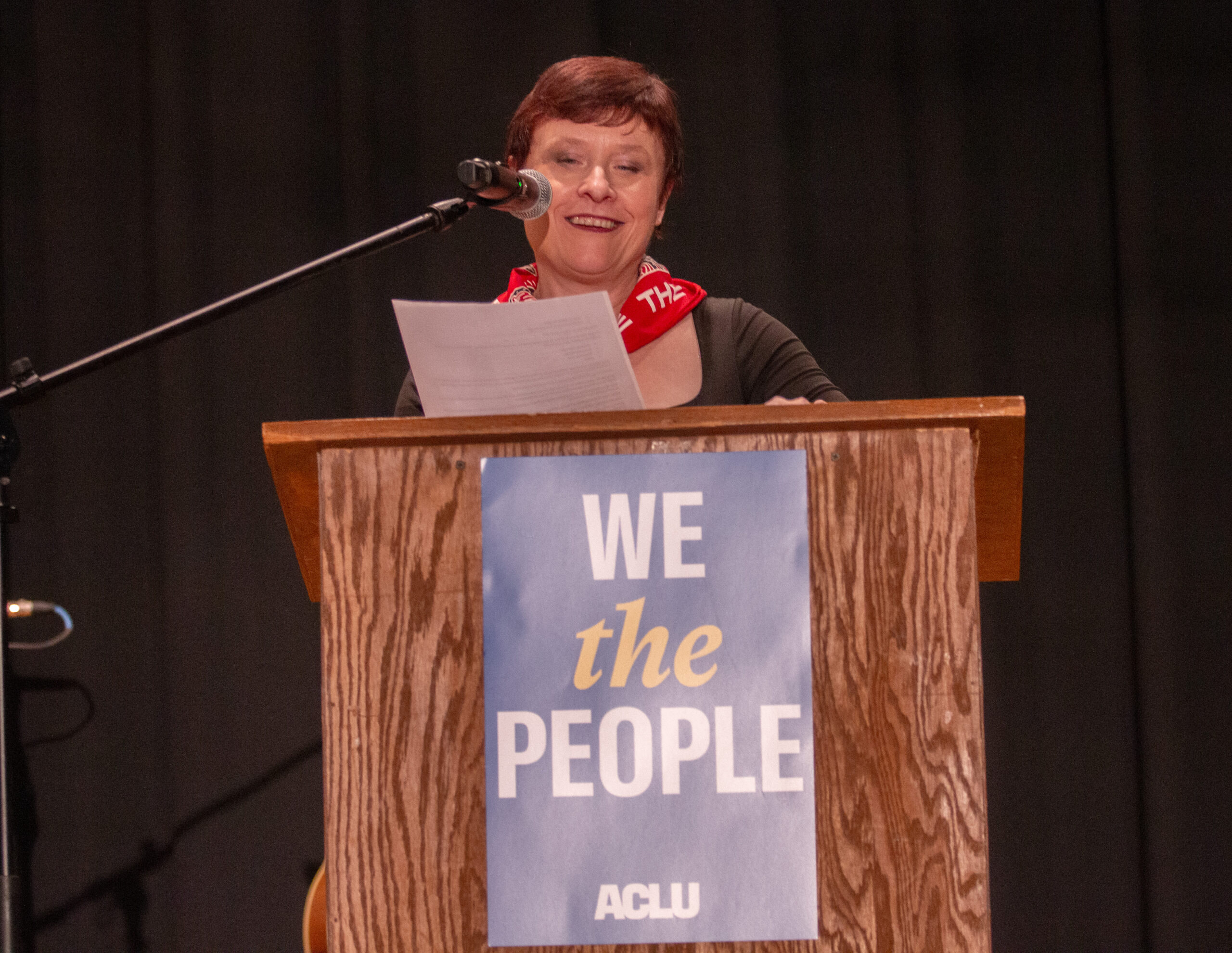 A person with short hair wearing a gray long-sleeve top and red "WE THE PEOPLE" bandana around their neck stands at a wooden podium, smiling while reading from papers. Microphone on stand positioned nearby. Podium displays blue and yellow "WE the PEOPLE" ACLU sign. Dark curtain backdrop.