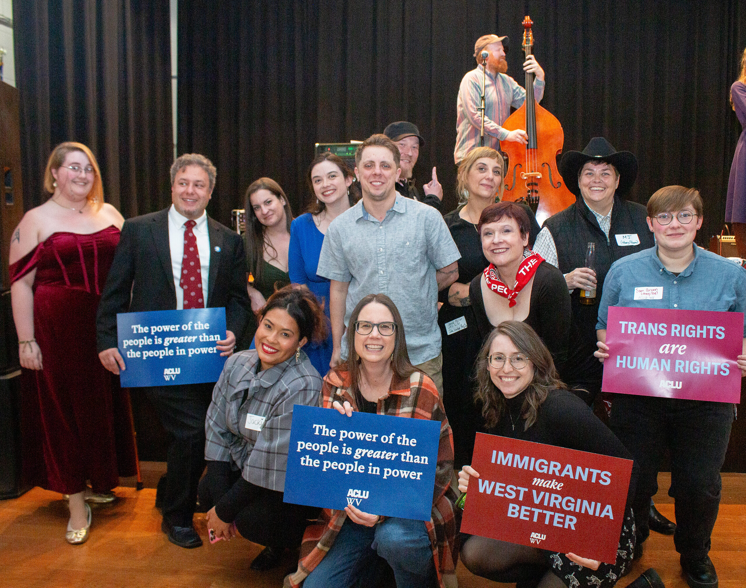 Large group photo on stage with musicians in background including upright bass player. Approximately 15 people arranged in two rows holding various ACLU advocacy signs including "The power of the people is greater than the people in power," "TRANS RIGHTS are HUMAN RIGHTS," and "IMMIGRANTS make WEST VIRGINIA BETTER." Mix of formal and casual attire with dark curtain backdrop.