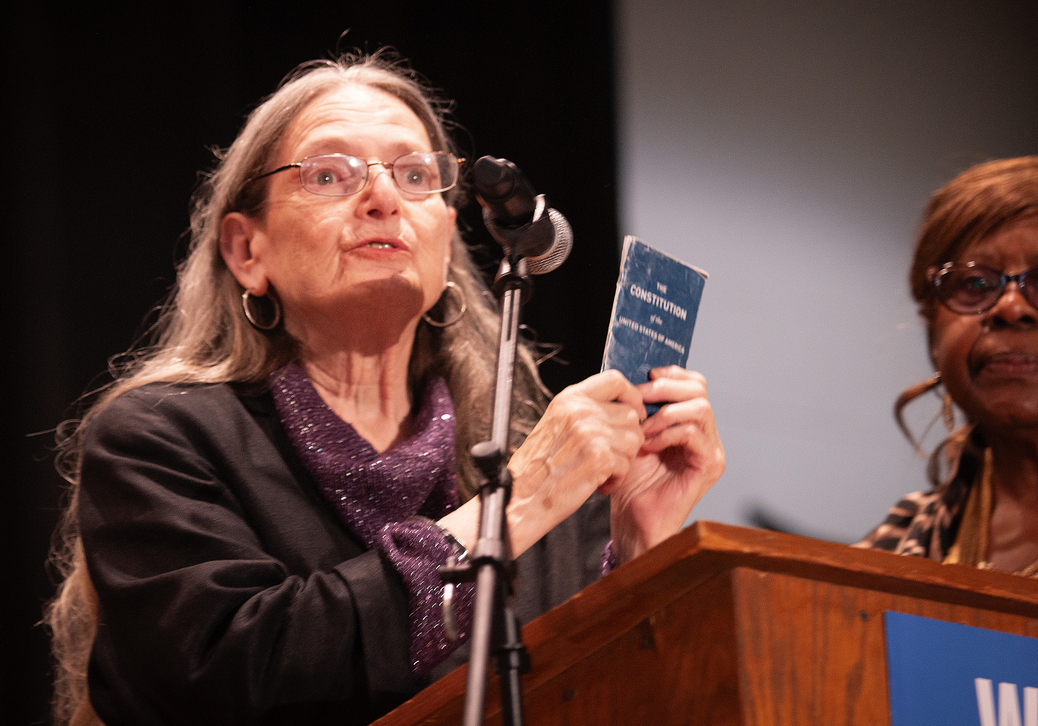An older person with long gray hair and glasses stands at a wooden podium speaking into a microphone, holding up a small blue pocket Constitution booklet in their raised hand. They wear a black jacket over a purple sparkly top. Another person partially visible in background to right. Podium appears to have ACLU signage.
