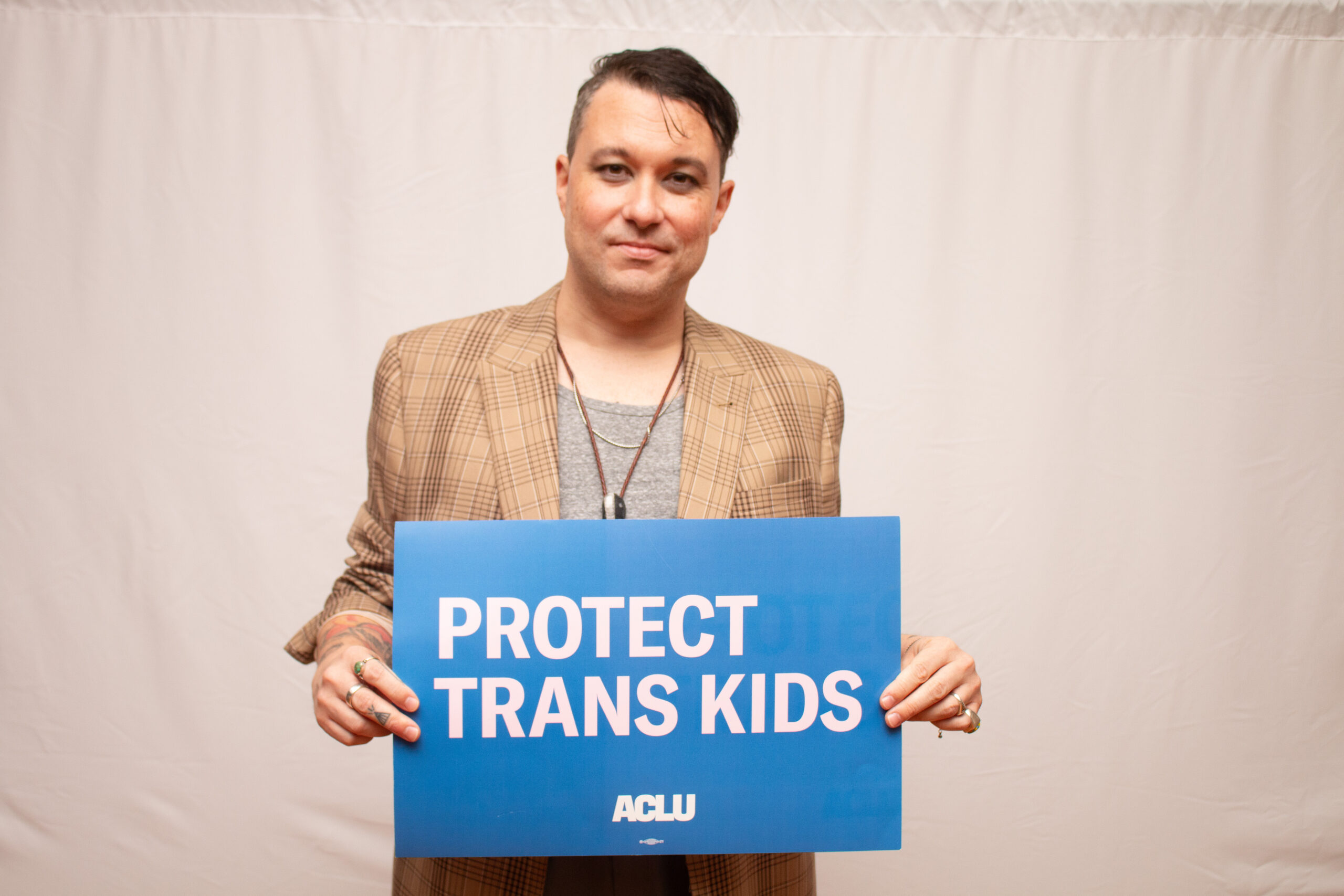 A person wearing a tan plaid blazer, gray t-shirt, and leather cord necklace holds a blue ACLU sign reading "PROTECT TRANS KIDS" against a light gray backdrop, looking directly at the camera with a serious expression.