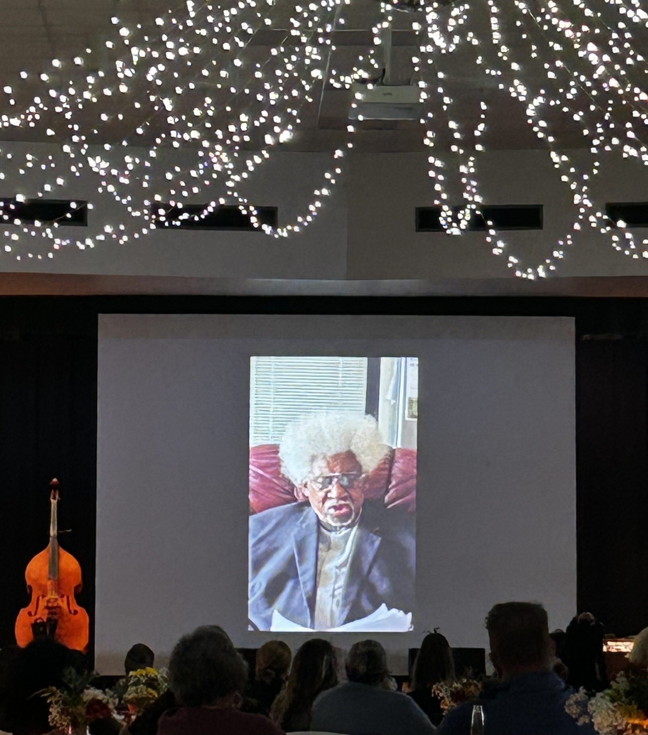 View from audience perspective showing large projection screen displaying video call with an elderly person with white hair, glasses, and blue cardigan seated in a home setting with pink bedding and window blinds visible. String lights hang overhead in the event space. Silhouettes of audience members' heads visible in foreground, with upright bass on left side of stage.RetryClaude can make mistakes. Please double-check responses.