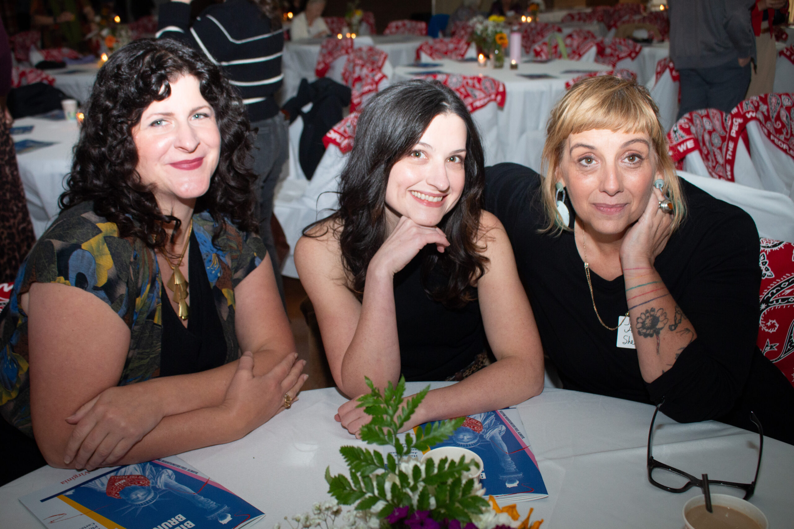 Three people sit at a white-clothed table at what appears to be a banquet or formal event. Tables with red patterned fabric decorations are visible in the background, along with other attendees. The three individuals, all wearing black tops, smile at the camera with candles and programs on the table.