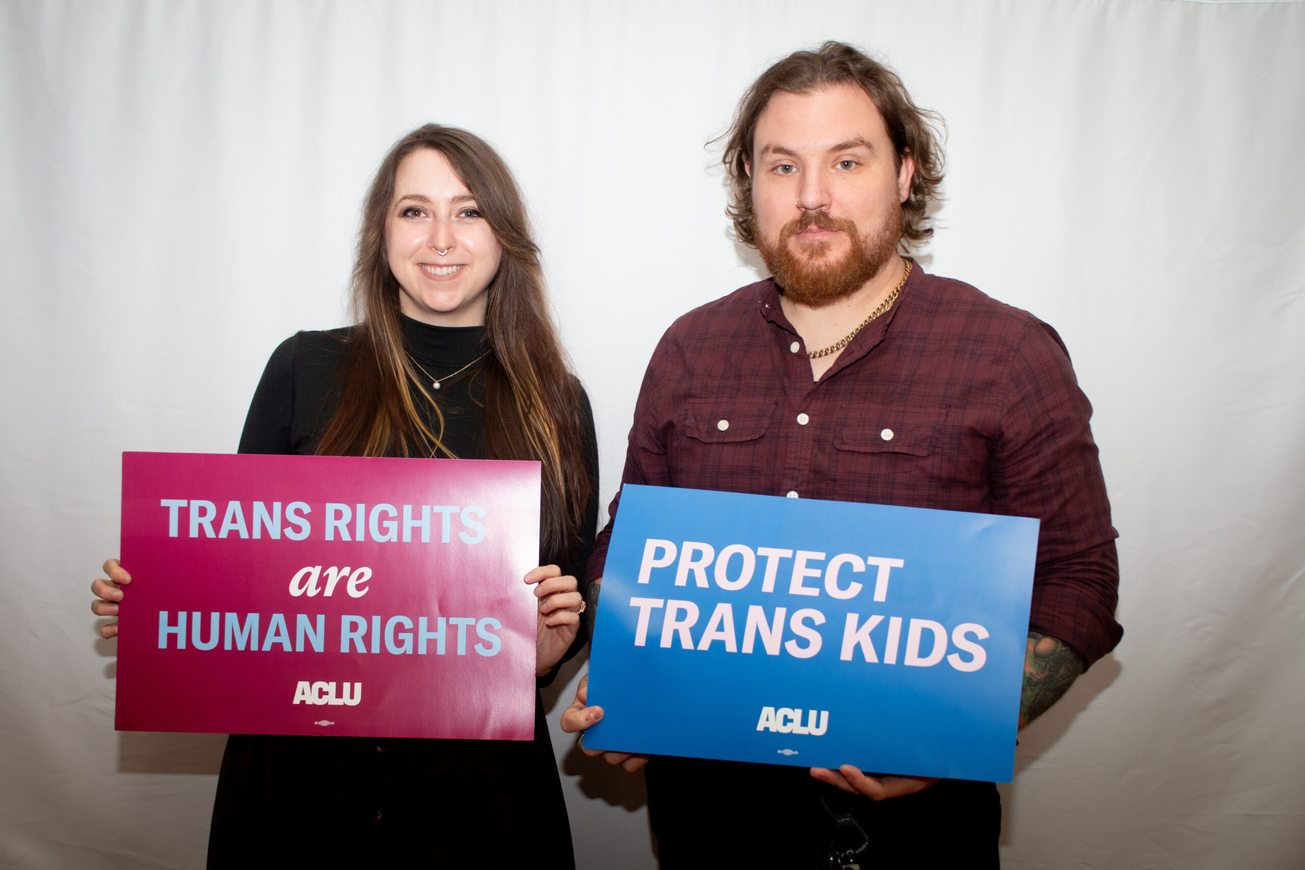 Two people hold advocacy signs against a white backdrop. The person on left in black top holds a pink ACLU sign reading "TRANS RIGHTS are HUMAN RIGHTS," while the person on right in burgundy button-up shirt holds a blue ACLU sign reading "PROTECT TRANS KIDS.