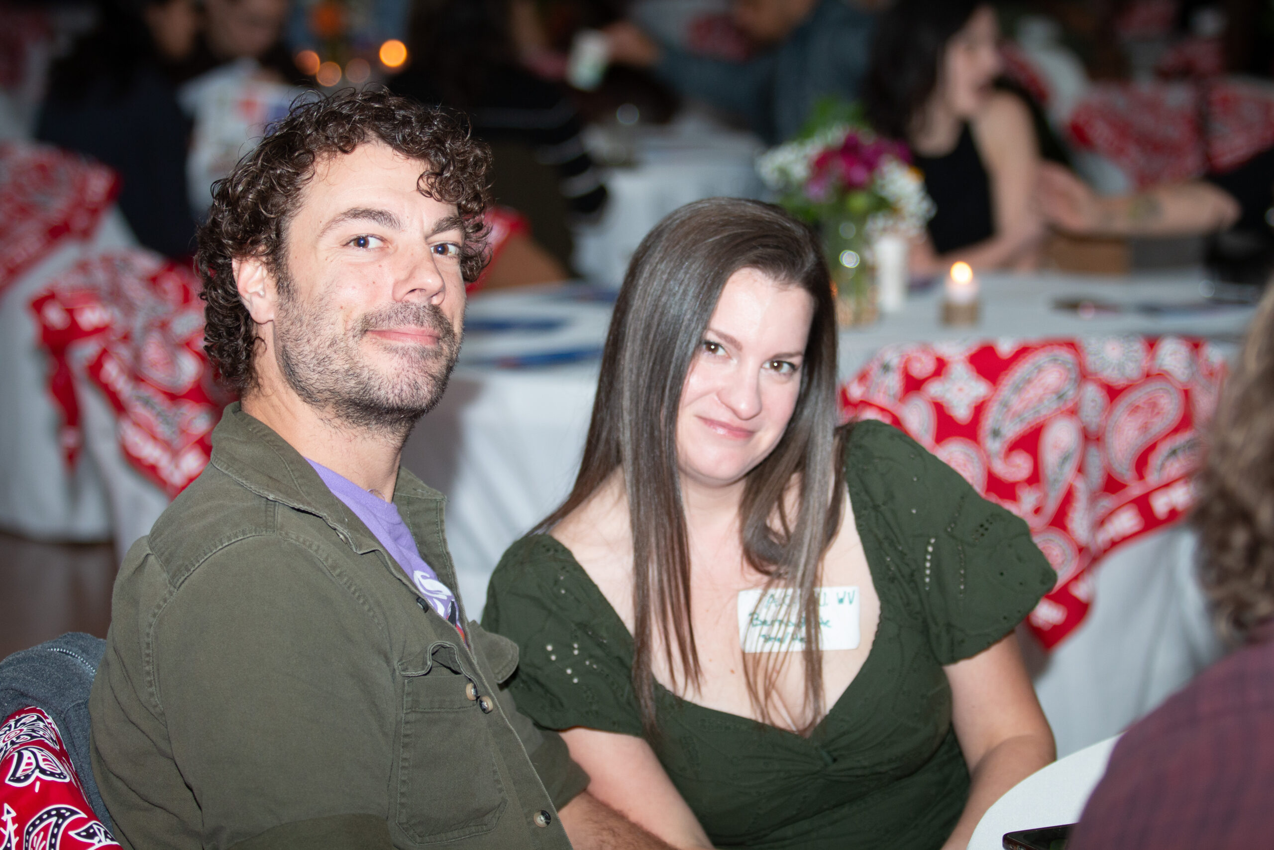 Two people pose together at an evening event with decorated tables visible in the background. The person on left wears an olive button-up shirt, and the person on right wears a dark green knit top with a name tag. Red patterned decorations and small floral centerpieces are visible on tables behind them.