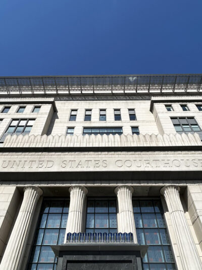 Modern United States Courthouse photographed from ground level against a clear blue sky. The imposing white stone building features classical fluted columns at the entrance, symmetrical rows of windows, and a distinctive solar panel canopy on the rooftop. The architectural design blends neoclassical elements with contemporary sustainable features, exemplifying modern federal architecture.