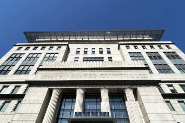 Modern United States Courthouse photographed from ground level against a clear blue sky. The imposing white stone building features classical fluted columns at the entrance, symmetrical rows of windows, and a distinctive solar panel canopy on the rooftop. The architectural design blends neoclassical elements with contemporary sustainable features, exemplifying modern federal architecture.