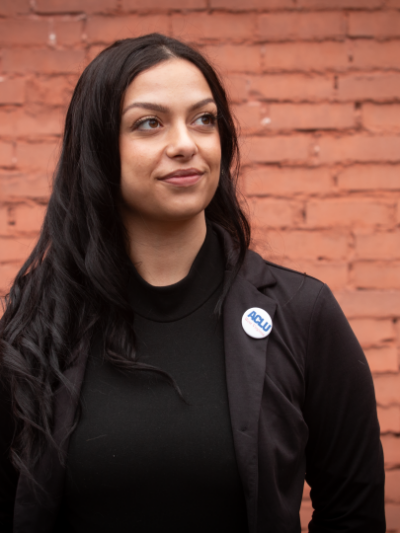 Portrait of a woman with long dark hair wearing a black blazer and turtleneck with a small ACLU West Virginia button pin, standing against a red brick wall background