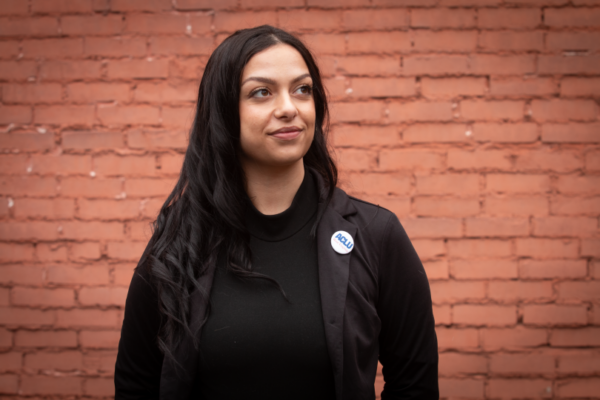 Portrait of a woman with long dark hair wearing a black blazer and turtleneck with a small ACLU West Virginia button pin, standing against a red brick wall background