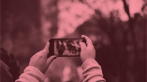 Someone holds up a smartphone to record or photograph a scene where a crowd of people has gathered. The image has a reddish-pink tonal filter applied to it. The person filming is in the foreground, their hands and device in focus, while the crowd behind them appears blurred. The scene suggests bystander documentation of a public event or gathering, capturing the modern phenomenon of people recording moments through their devices rather than simply observing directly.